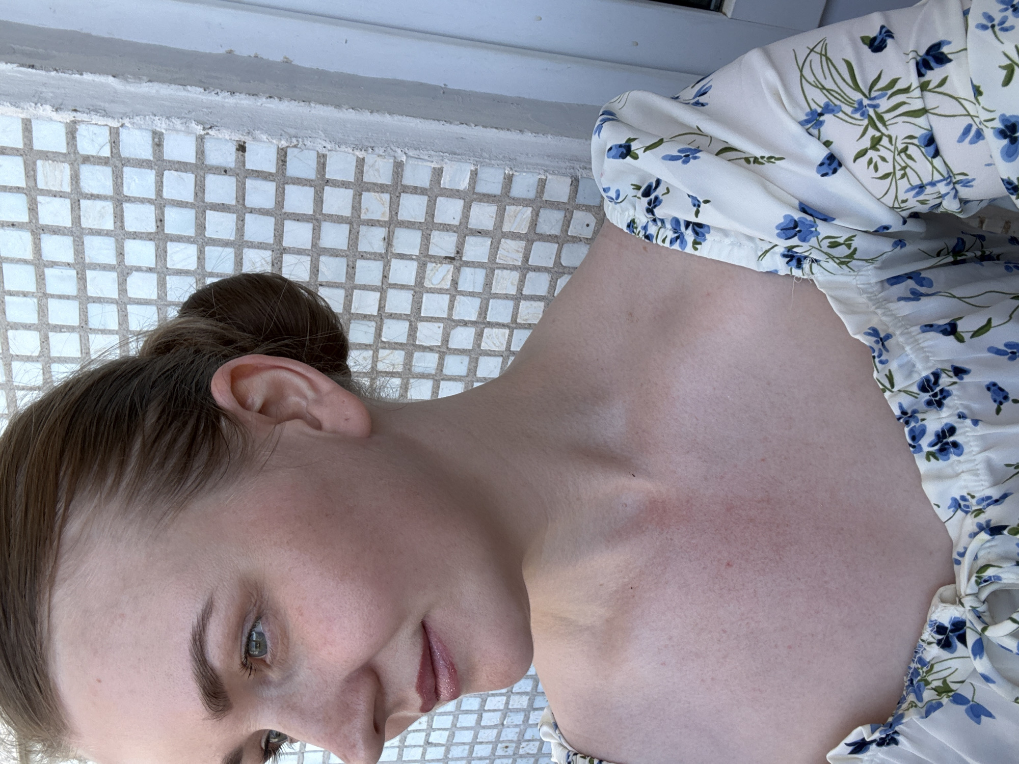 A woman is photographed at close range. She has her hair pulled back in a bun and is wearing a white blouse with a blue floral pattern. Her skin is fair and she has light-colored eyes. The background consists of a tiled surface and a white painted structure. The location is specified as Chaikovsky, Russia.
