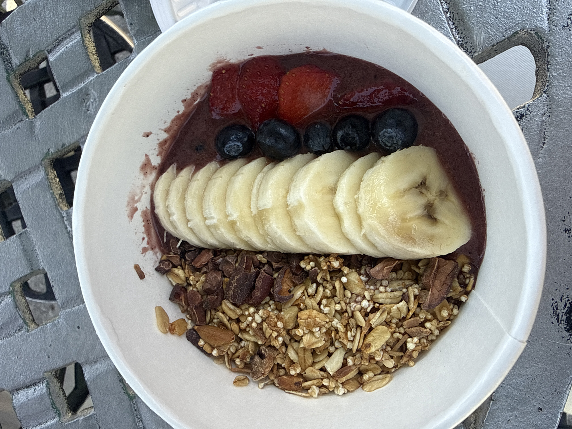 The image shows an acai bowl in a white paper bowl, likely photographed outdoors in daylight. The bowl contains a dark purple acai base topped with sliced strawberries, blueberries, sliced bananas, and granola. It's resting on a grey surface with a decorative, possibly woven, pattern. The location is San Andrés Cholula, Mexico.