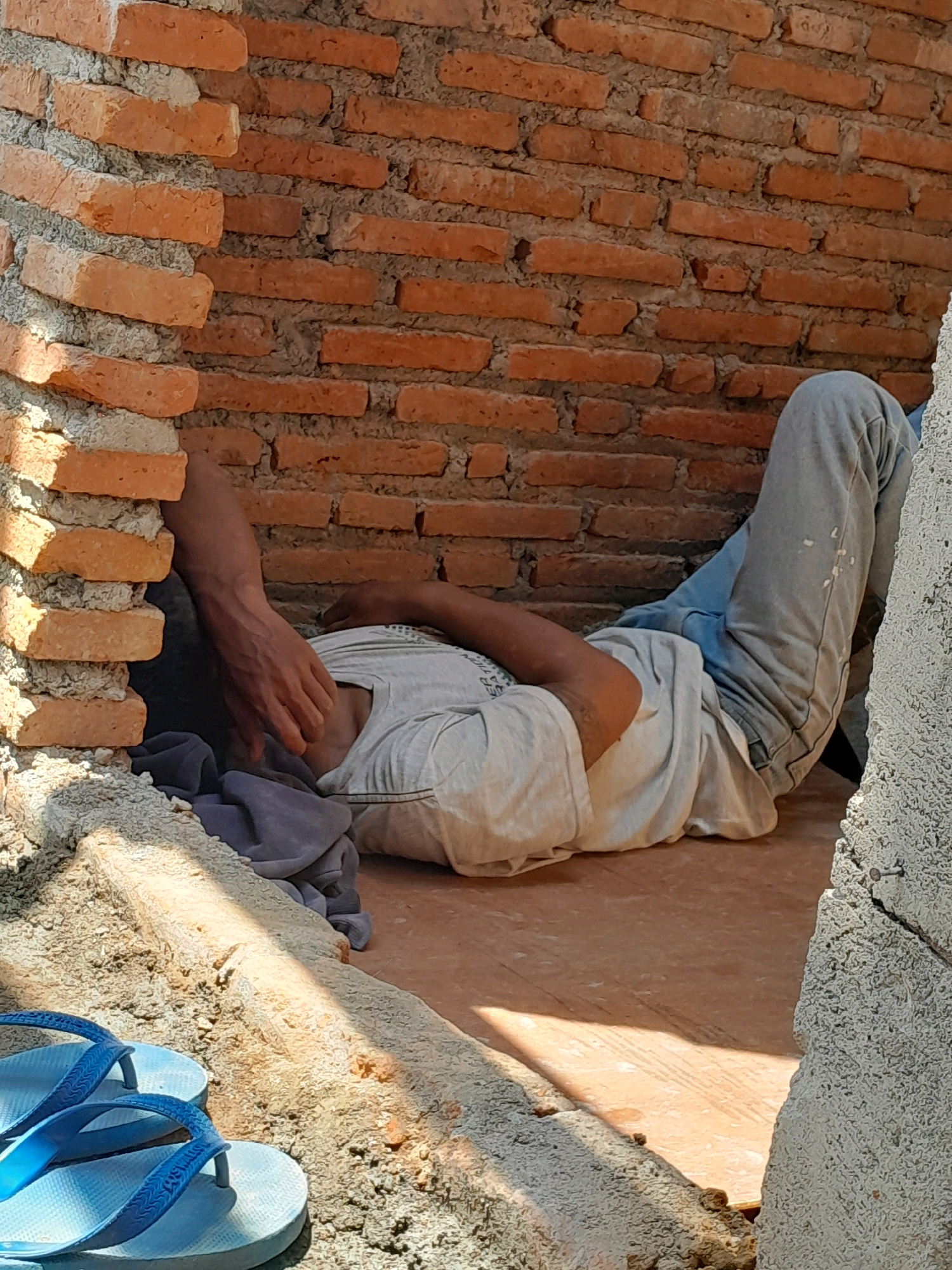 A person is lying down on their side, propped against a brick wall. Their head is obscured by their arm and a dark gray cloth. They are wearing a light-colored t-shirt and light blue jeans. The scene appears to be an outdoor construction area, with unfinished brickwork and piles of sand or gravel visible. A pair of blue flip-flops are in the foreground. The sunlight casts strong shadows across the ground. The location is identified as Karangduren, Indonesia.