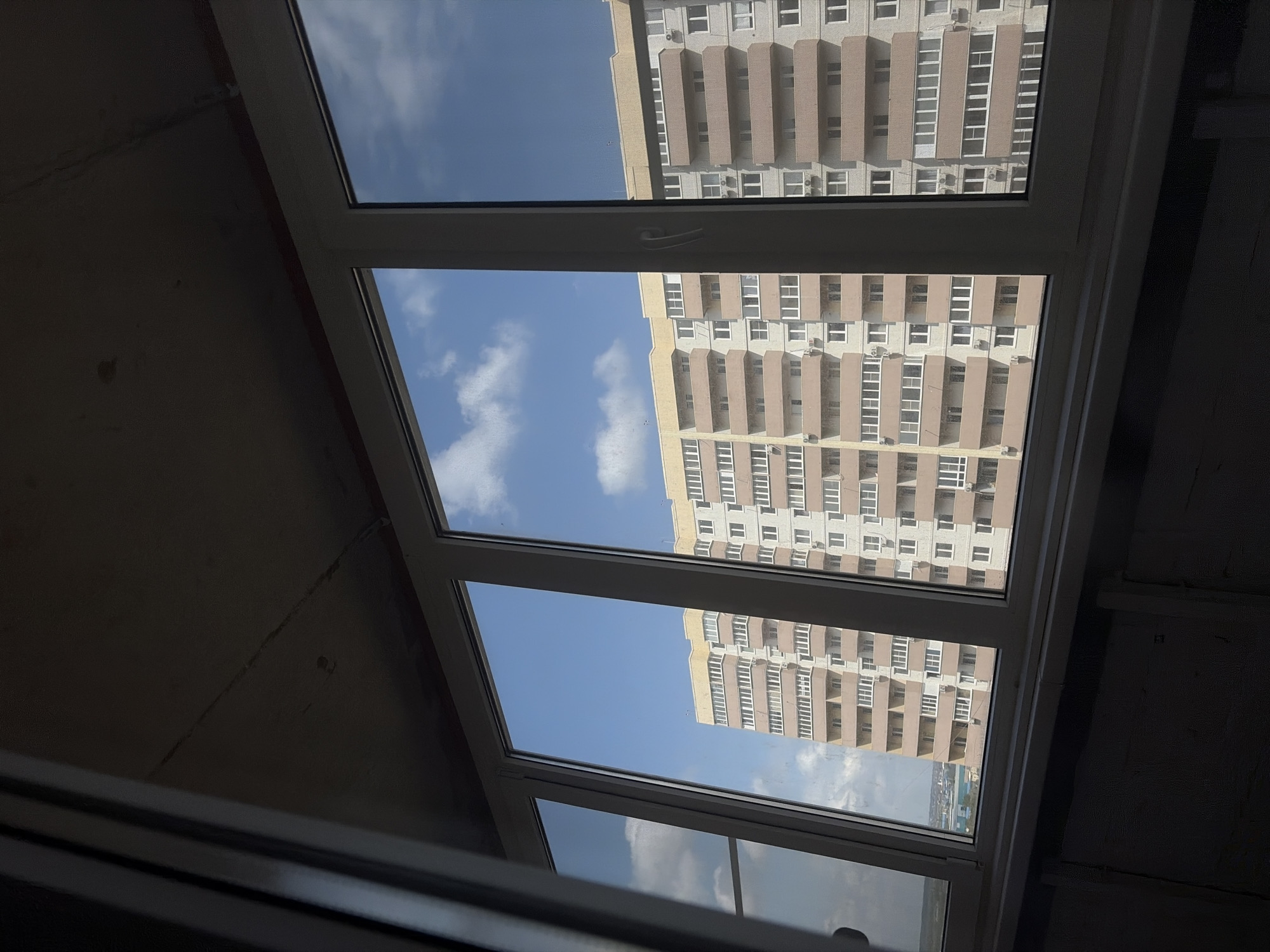 The image shows a view from inside a building, looking out through a multi-pane window. The window frames are white, and the view is of a clear blue sky with some scattered white clouds, as well as a tall apartment building across the street. The apartment building is light beige with rows of balconies. The interior of the room is dimly lit, with gray walls and the window taking up a large portion of the frame.