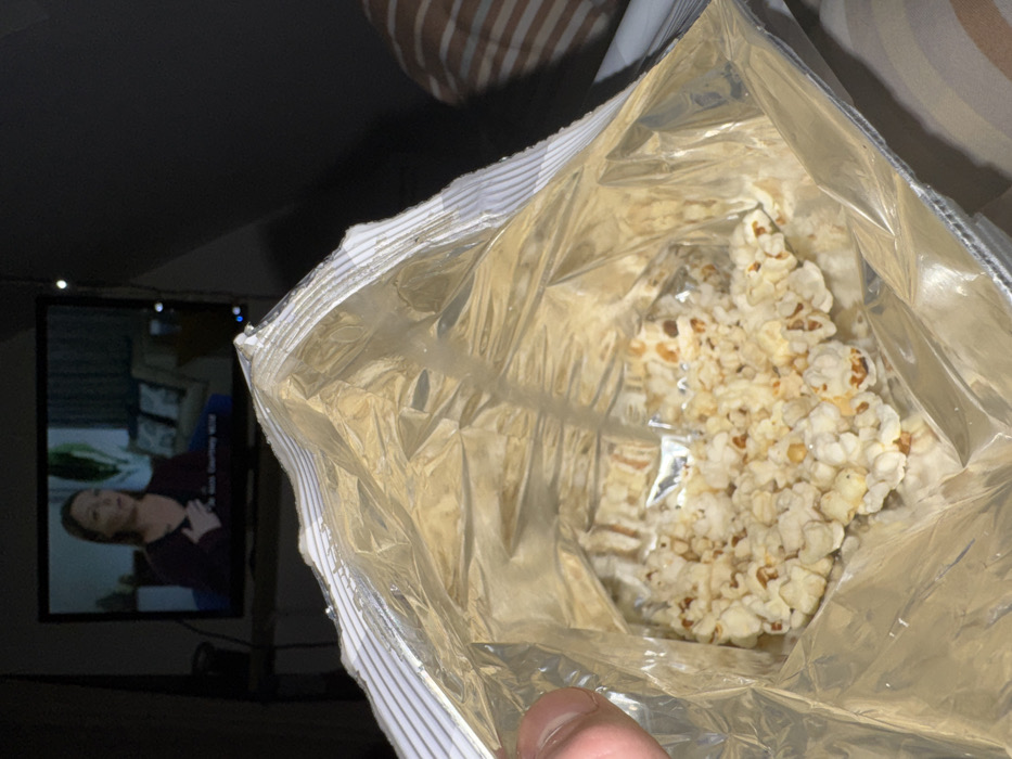 An opened bag of popcorn reveals a mostly empty interior filled with scattered white and lightly browned popcorn. A television screen in the background shows a woman in a scene, partially visible in the dark room. The bag has a metallic interior and a striped edge. The scene suggests a relaxed indoor setting, possibly enjoying a snack while watching TV. A finger is visible in the bottom of the image.
