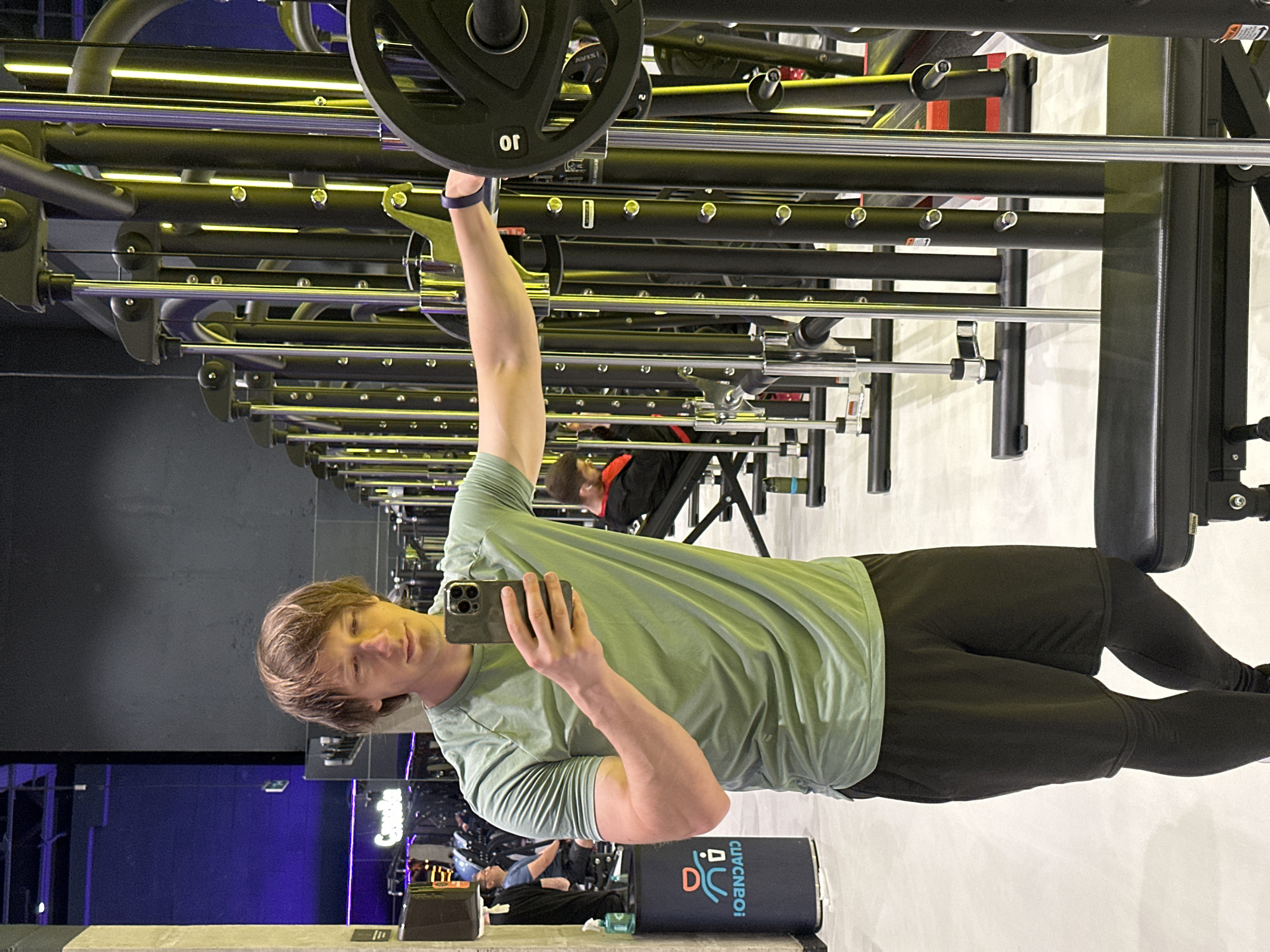 A young man in a light green long-sleeved shirt and black pants is taking a selfie in a gym. He is holding his phone in his right hand, which is raised and pointing towards a mirror. His left arm is also raised, holding a weight plate on a barbell. The barbell is part of a squat rack, and other gym equipment, including more barbells and weight plates, are visible in the background. The gym appears to be modern, with black and silver equipment and some yellow lighting. In the background, another person can be seen working out. The overall mood is one of fitness and self-care. The text "CLVCNEBOI" is visible on a blue cylindrical object in the foreground.