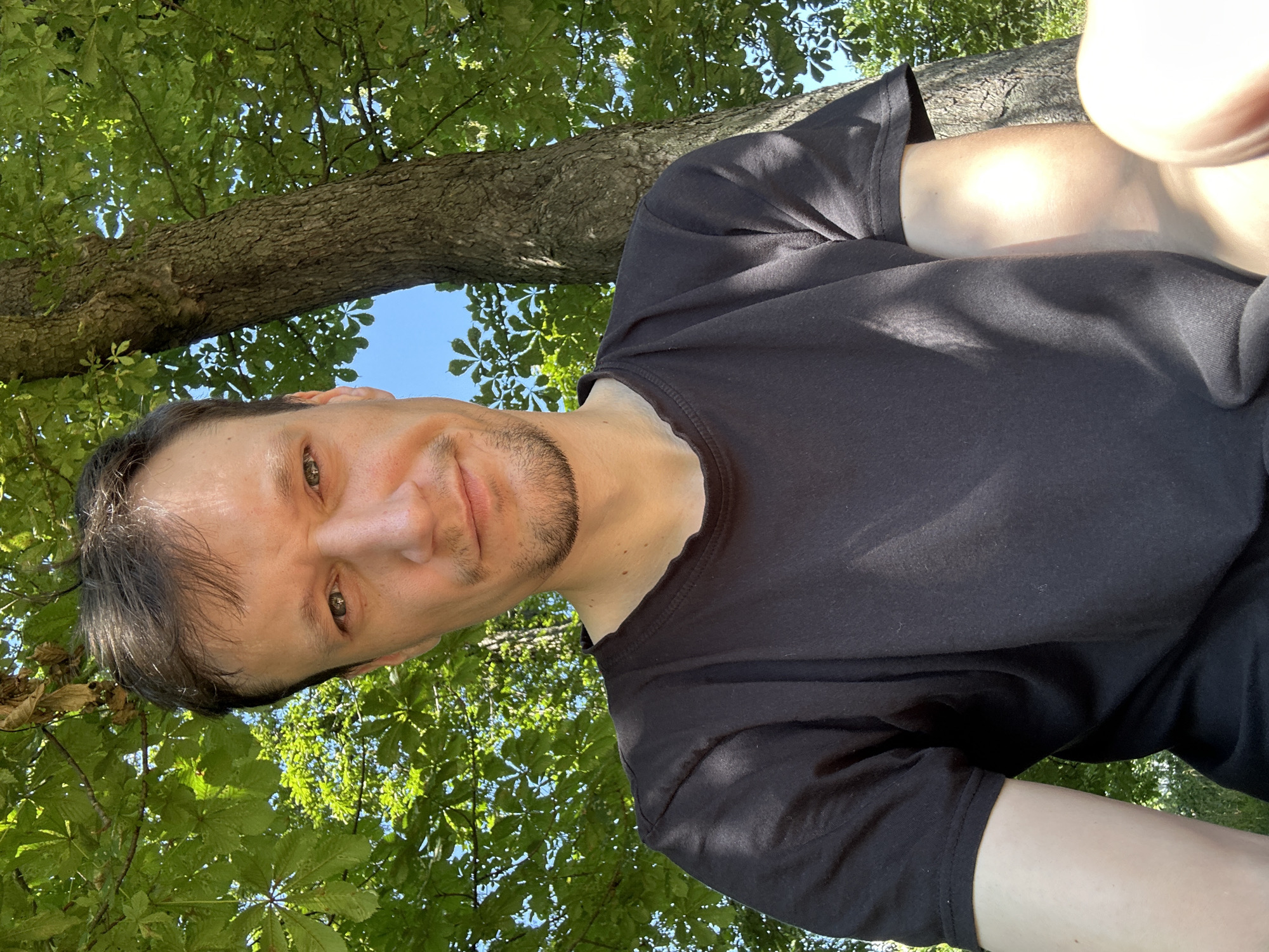 A medium shot photo, taken outdoors on a bright, sunny day in Makiv, Ukraine, features a man looking directly at the camera with a slight smile. He is wearing a black t-shirt. The man is positioned in front of a large tree with green leaves and a clear blue sky visible behind it. The overall scene suggests a casual, outdoor setting.