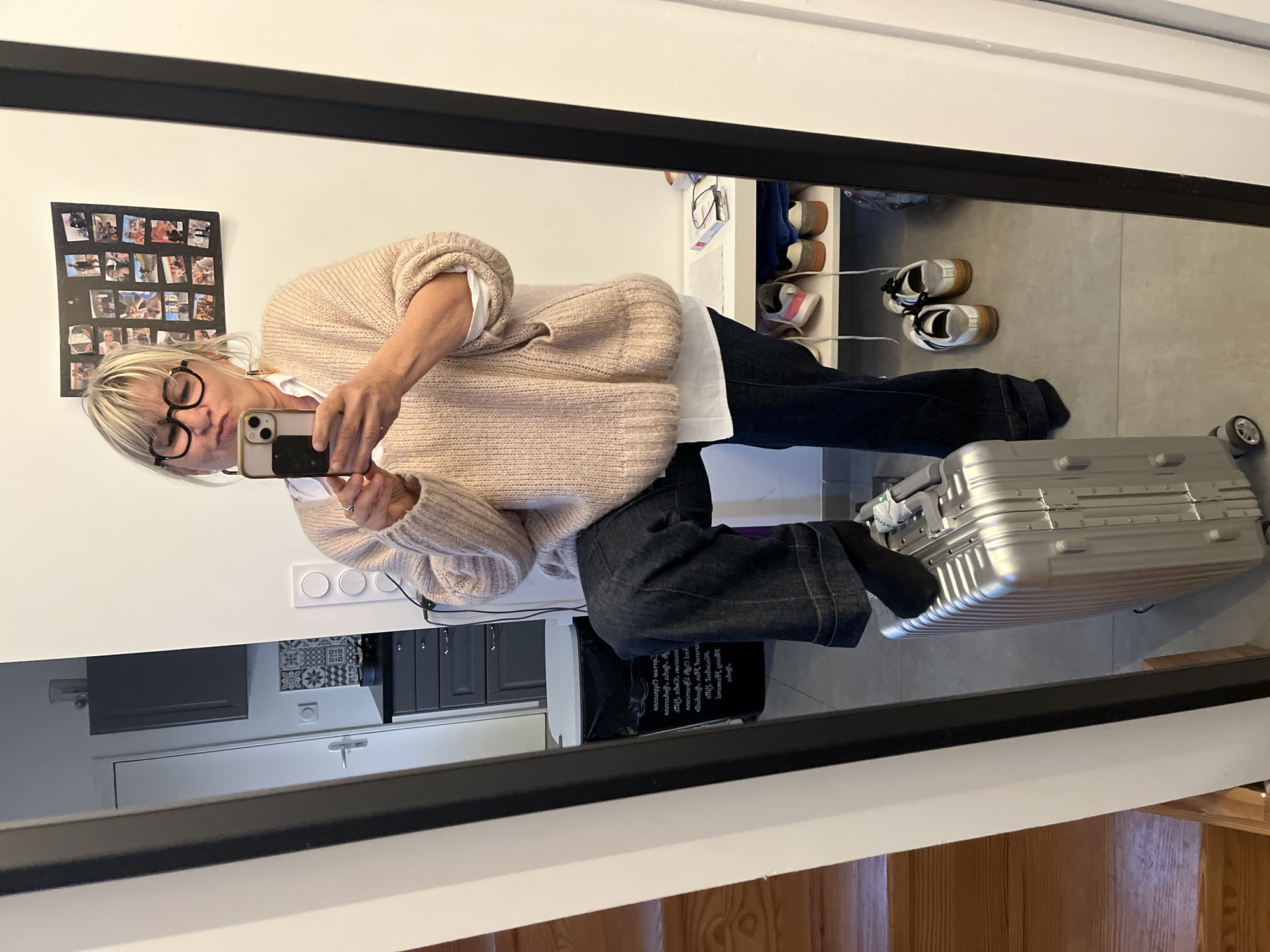 A woman takes a mirror selfie indoors. The reflection shows her wearing a blush-toned sweater over a white shirt, dark jeans, and black socks, with round glasses and blonde bangs. She is holding a smartphone in her right hand to take the picture. Beside her is a silver suitcase.
Behind her is a white wall with a collage of photos arranged in a grid. There are light switches on the wall. Lower in the reflection, the mirror reveals a portion of a kitchen with grey cabinets and an open drawer, plus a black bag.
To the right of the woman, there is a shoe rack holding several pairs of shoes. The floor is tiled in a light grey color. In the bottom right, wooden stairs are visible, indicating the presence of a staircase.
The location is Chambœuf, France.