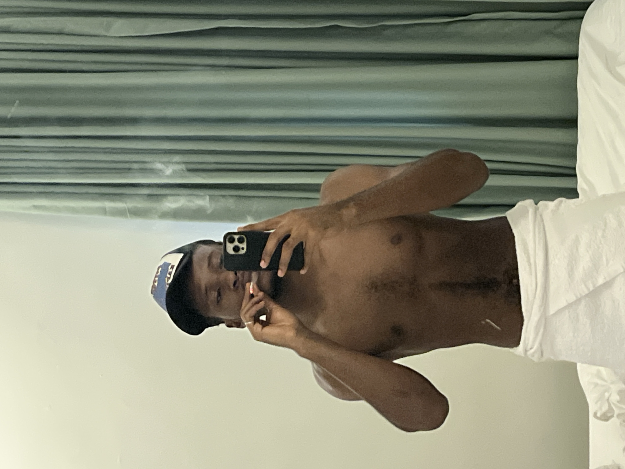A mirror selfie captures a young, dark-skinned man in an indoor setting in Gwarinpa, Nigeria. The image is rotated 90 degrees clockwise, showing the man lying horizontally. He is shirtless, with a white towel wrapped around his waist, covering his lower body. He wears a blue and white baseball cap.
The man holds a black smartphone, likely an iPhone due to its distinct camera array, in his right hand, pointing it towards the mirror to take the photograph. His left hand is raised towards his mouth, holding a small, white cylindrical object, possibly a cigarette or vape. A faint wisp of smoke or vapor is visible in the upper left background, suggesting the object is lit. A silver-colored ring is noticeable on his left ring finger. There are a few light-colored patches or marks on his right arm.
The background consists of horizontally striped teal or greenish-grey curtains in the upper portion of the frame, and a plain light-colored wall or surface in the lower portion. A white pillow or bedding is visible on the far right side of the image. The scene suggests a private room, possibly a bedroom or bathroom. The lighting appears to be from an indoor source. No discernible text is visible on the man's cap or elsewhere in the image.
