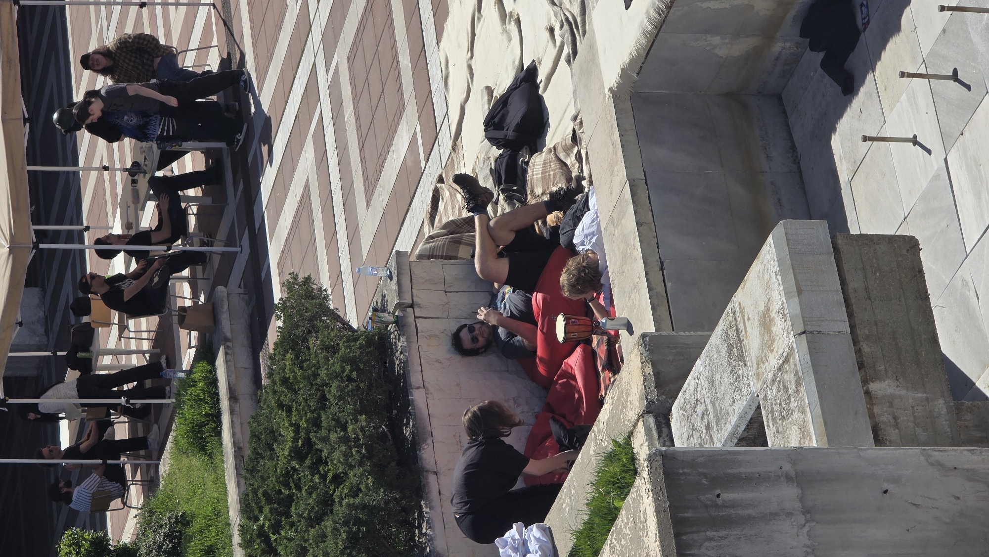 The image depicts an outdoor scene, possibly taken from an elevated position.
On the left side, several individuals are seated in chairs under a white canopy, possibly a tent. The background shows a building with a striped pattern.
On the right side, another group of people are gathered in what appears to be a concrete structure, possibly a fountain or a raised seating area. They are sitting on blankets and pillows. One person is lying down with their legs up, another is playing a small drum, and another is sitting.
The presence of greenery, including a bush, suggests this is an urban outdoor setting. Based on the prompt, the location is Blagoevgrad, Bulgaria.