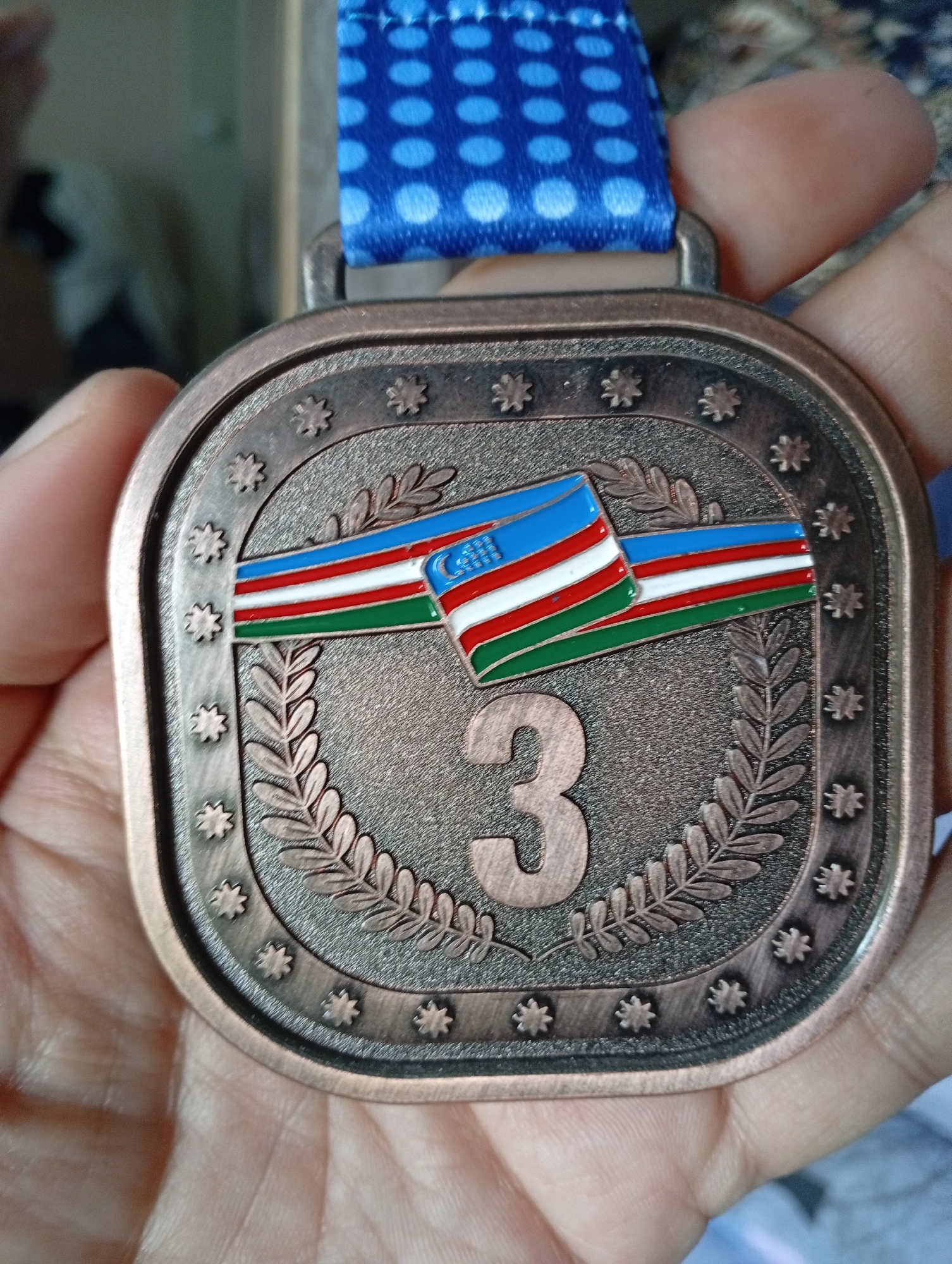 A close-up, eye-level shot shows a hand holding a bronze medal. The medal is square with rounded corners and has a patterned border of small star-like emblems. In the center, a stylized laurel wreath surrounds the number "3" in a raised, copper-toned font. A ribbon, featuring a blue background with a white polka-dot pattern, is attached to the top of the medal. A small flag, with horizontal stripes of blue, white, and green, and a crescent moon and stars in the canton, is depicted across the upper part of the laurel wreath. The background is out of focus, with hints of indoor elements and soft lighting. It is not possible to determine the location in Angren, Uzbekistan from the image alone.