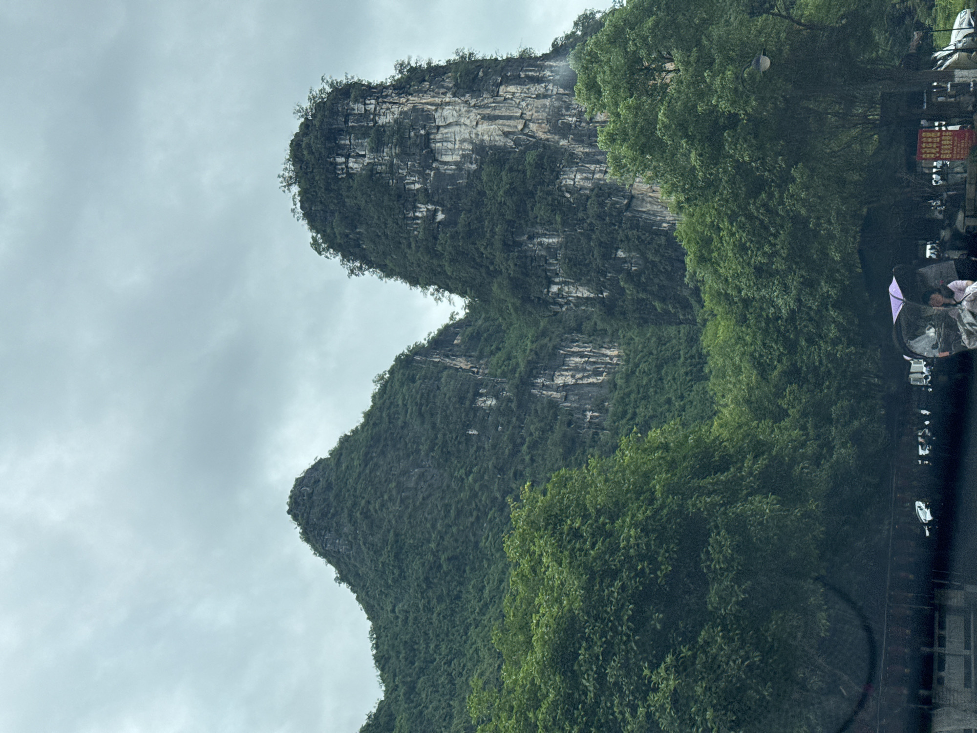 The image depicts a landscape featuring two prominent karst mountains in Guilin, China. The sky is overcast. The mountains are covered in lush greenery and show visible rock formations. To the right, there are trees and what appears to be a roadside scene, including people and structures.