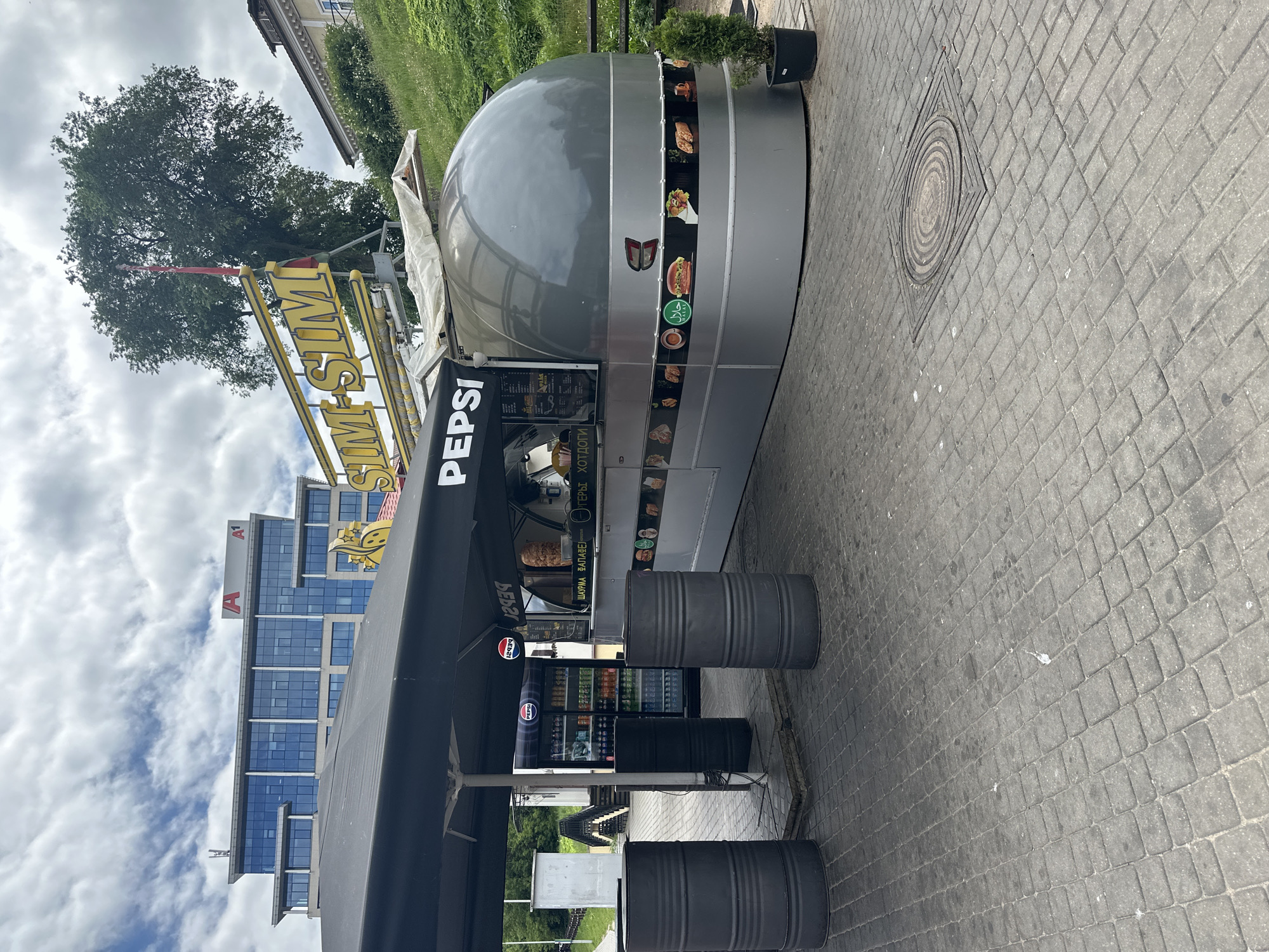 In an outdoor urban setting, likely a city square or pedestrian area, a contemporary fast-food kiosk named "SIM-SIM" is prominent. The kiosk is a modern, silver, capsule-shaped structure with a large black awning extending over a waiting area. The "SIM-SIM" branding is displayed in large, yellow, illuminated letters above the kiosk. A prominent "PEPSI" logo is visible on the awning.
In front of the kiosk, there are two large, dark grey cylindrical trash bins. To the left of the kiosk, a large glass-doored refrigerator stocked with various beverages, predominantly Pepsi products, is visible. The ground is paved with grey interlocking brick pavers.
Behind the kiosk, a modern office building with a glass facade and the red "A1" logo is visible, suggesting this is a commercial district. A large, mature tree stands behind the kiosk, and a grassy slope with a wooden fence runs along the right side of the image.
The kiosk's lower section features a band of images showcasing food items, including burgers and what appear to be fried chicken or pastries. A green "Halal" symbol is also visible on this band. A small potted evergreen tree is placed to the right of the kiosk.
The sky is overcast with scattered clouds. The scene appears to be captured on a daytime, possibly slightly humid, day. No people are visibly present in the immediate vicinity of the kiosk. The overall impression is of a functional, modern food service establishment in a well-maintained urban environment. The city of Minsk, Belarus, is indicated as the location.