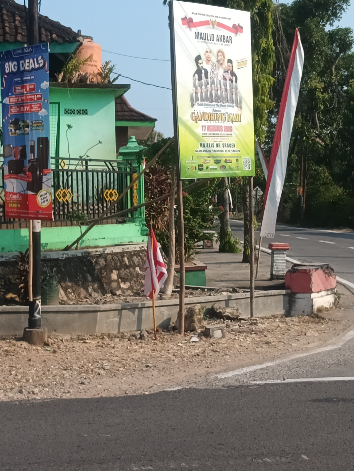 This is a photograph of a street scene in Palemrejo, Indonesia, on a clear day with bright sunlight. In the foreground, a paved road is visible, bordered by a gravelly shoulder and a low concrete wall. Behind the wall, a sidewalk area is present, with a black cylindrical pole supporting a banner. To the left, a green building with a dark tiled roof is partially visible, with some vegetation growing on top.
Dominating the right side of the image is a large, vertical poster advertising an event titled "MAULID AKBAR GANDRUNG NABI". The poster features images of several people, including what appears to be a religious leader, and a group of men in traditional attire. Below the main title, it states "17 AGUSTUS 2023". Further down, it lists "MAJELIS NR SRAGEN". There are also various logos and what seems to be a QR code at the bottom of the poster.
To the right of this poster, an Indonesian flag, red and white, is displayed vertically. Behind the posters and the building, lush green trees form a dense backdrop. The environment suggests a semi-rural or suburban setting, with power lines and utility poles also visible. The overall impression is one of an outdoor public space with a promotional event being advertised.