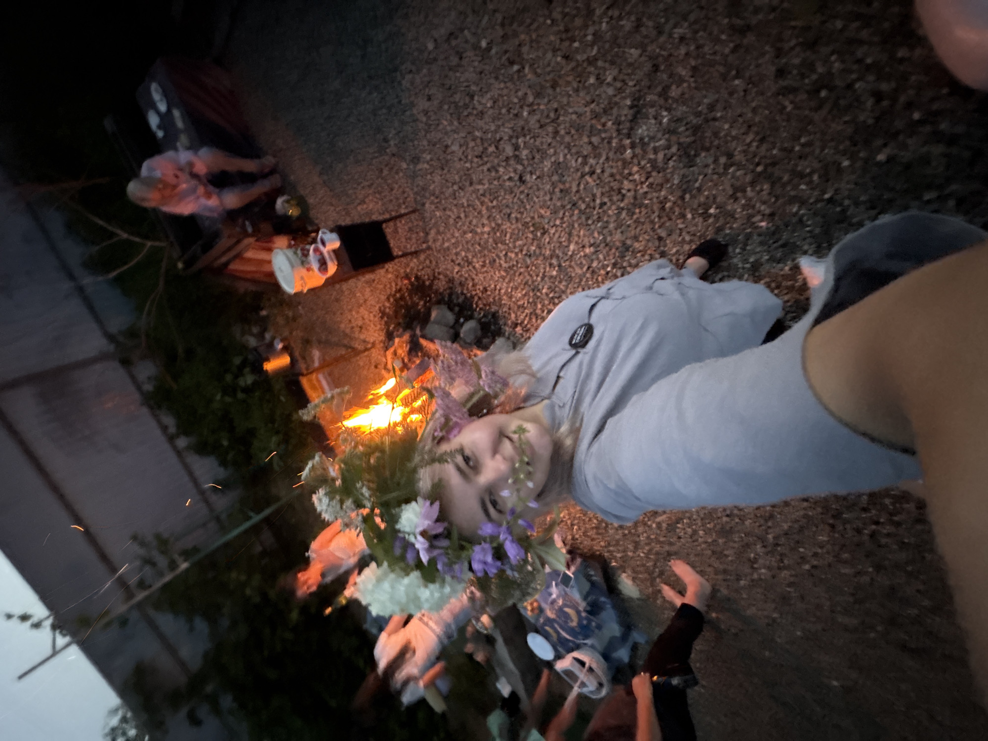 A young woman wearing a flower crown is taking a selfie at night. She is smiling and looking at the camera. In the background, a bonfire is burning, and several people are gathered around it. The scene appears to be in a backyard or park. The woman is wearing a light-colored shirt. The ground is covered in gravel. There are some trees and plants in the background, as well as a fence. The lighting is dim, with the bonfire providing the main source of illumination. The woman is in the foreground, and the other people and objects are in the background, out of focus.