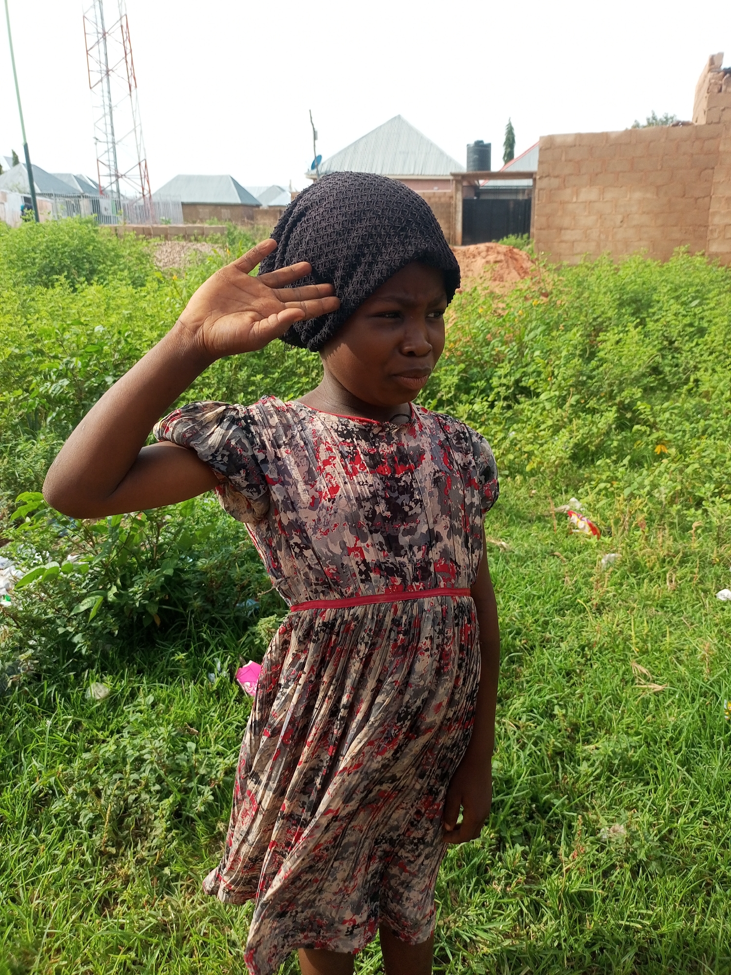 A young Black girl, wearing a dark knit beanie and a printed dress with red trim, stands in an outdoor, grassy area. She is facing the right side of the frame, with her left arm raised and hand covering her eyes. In the background, several buildings are visible, including a brick structure and a telecommunications tower. The area appears to be in a rural or semi-urban setting, with abundant green vegetation.