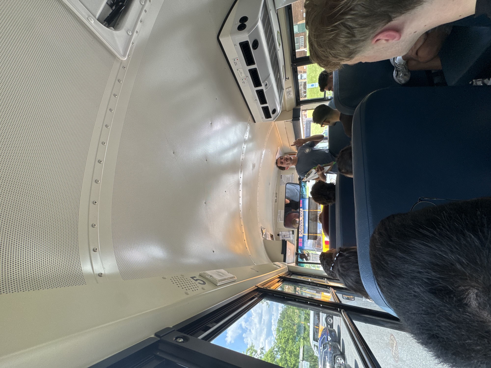 The image shows the interior of a yellow school bus, viewed from a rear seat looking forward. A young man in a grey t-shirt is standing near the front of the bus, holding a clipboard and gesturing with his left hand. He appears to be addressing the passengers. Several passengers are visible in their seats, facing forward. Outside the bus windows, trees and a cloudy sky are visible, along with other vehicles on a road. The bus is equipped with overhead air vents and a ceiling panel with perforated holes. The overall scene suggests a group embarking on or disembarking from a bus journey, possibly a field trip or transportation to an event. The location is identified as Washington Township, United States of America.