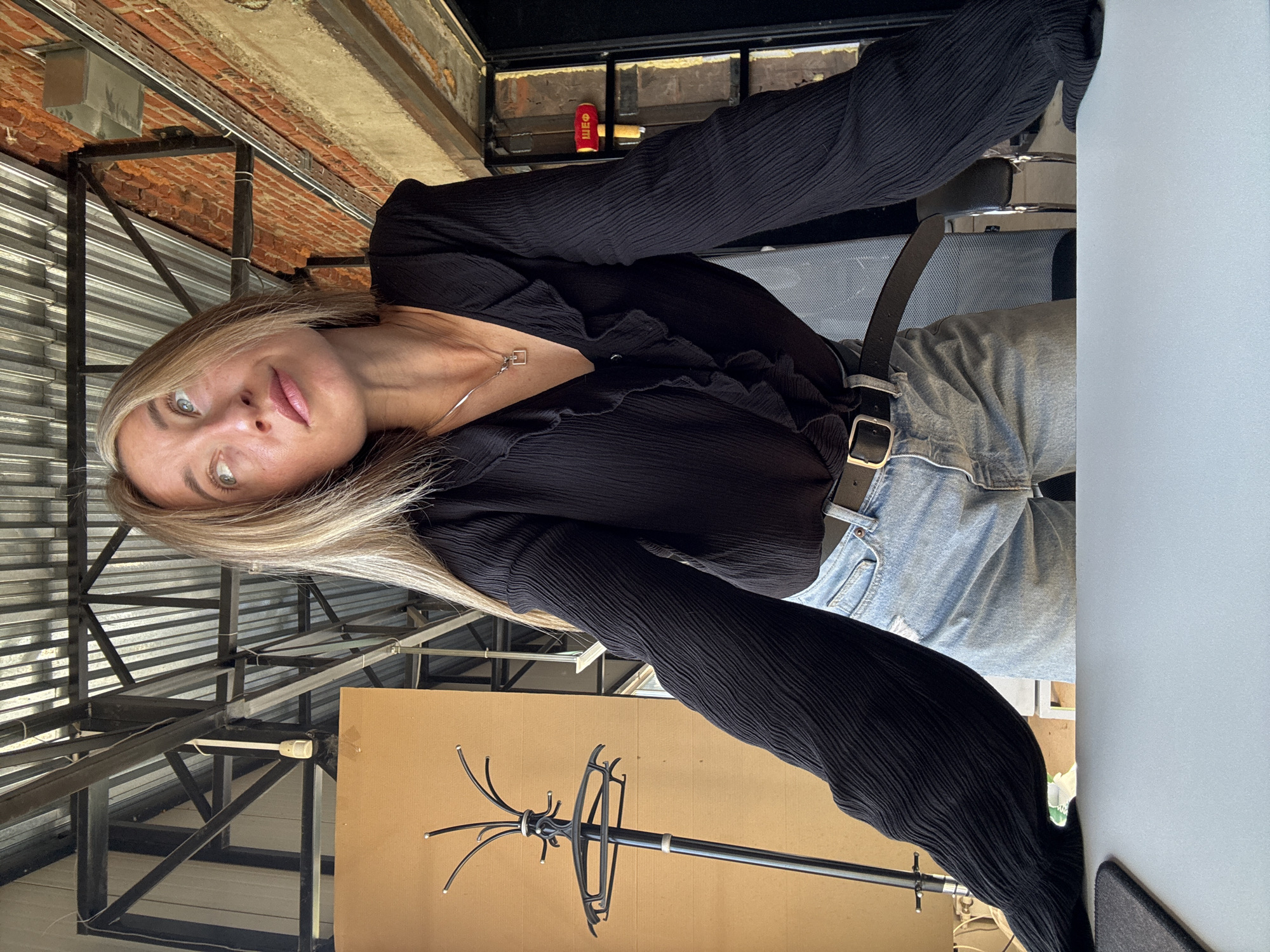 The image shows a young woman with blonde hair, wearing a black ruffled blouse and blue jeans, leaning over a desk. She is looking upwards and to the right. The background suggests an indoor, possibly industrial or loft-style space, with exposed metal beams and corrugated metal roofing. A brick wall is visible to the right. To the left, a coat rack with several hangers stands near a wooden panel. The scene is well-lit, likely by natural light, creating a bright atmosphere. The context of the city of Chaikovsky, Russia, is not directly evident from the visual elements within the image itself.