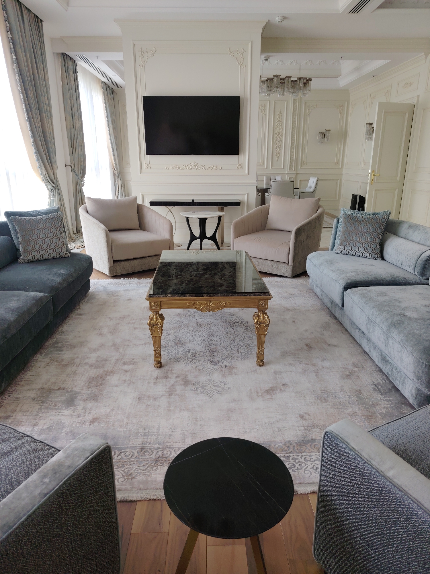 The image depicts a luxurious living room. A large, light-colored rug with an intricate pattern covers the floor. In the center of the room is a rectangular coffee table with a dark marble top and ornate gold legs. Two light beige armchairs are positioned on either side of the coffee table. Along the perimeter of the room, there are two long, light blue sofas. A large, black flat-screen television is mounted on the wall. The room features ornate white molding and decorative wall panels, along with elegant curtains and a chandelier in the background. The overall aesthetic suggests a sophisticated and modern design. There are no people visible in the image. The location is not specified in the image.