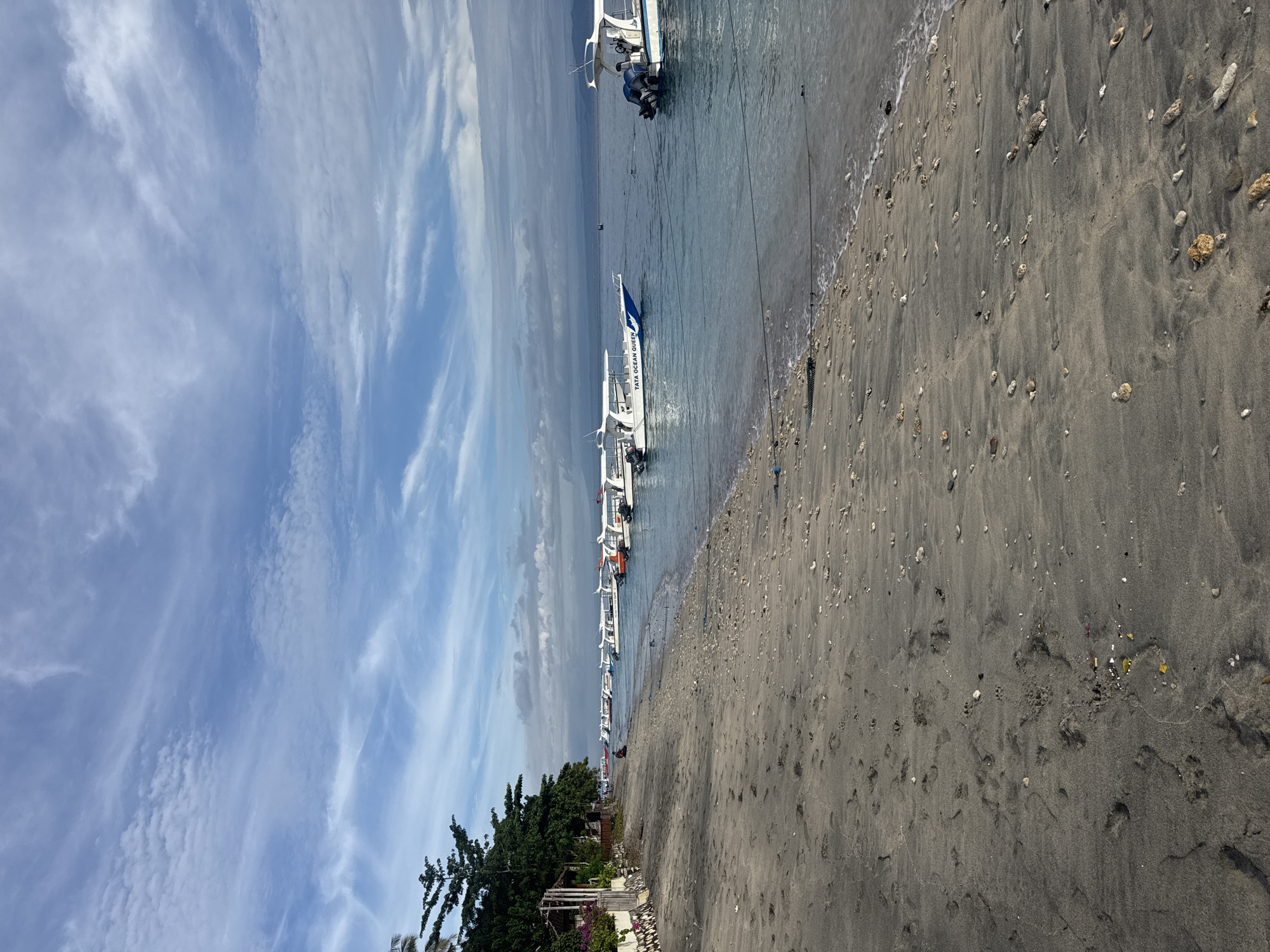 The image depicts a beach scene in Nusa Penida, Indonesia. The beach consists of dark grey sand dotted with small pebbles and coral fragments. Several white boats are moored close to the shore, aligned parallel to the beach. A few small buildings and trees are visible near the shoreline. The sky above displays a mix of blue and wispy white clouds. No people are clearly visible in the image.