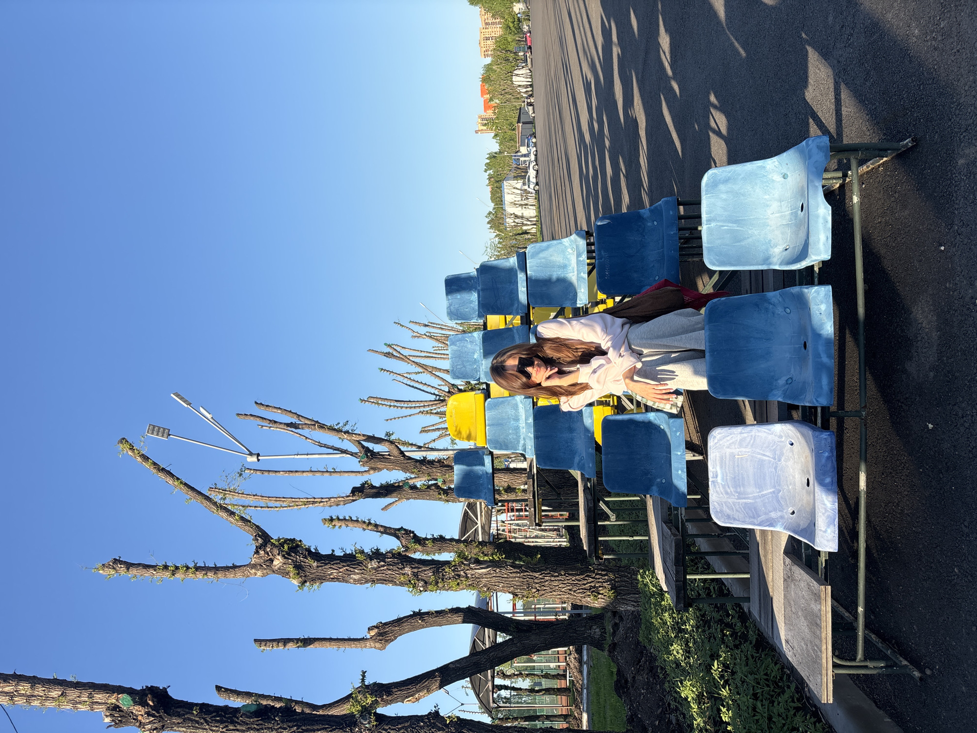 A medium-wide shot of a seated woman with long brown hair wearing sunglasses, a light pink sweatshirt, and gray sweatpants. She is positioned across several blue and yellow plastic stadium-style seats arranged in rows. The seating area is adjacent to a tree with trimmed branches. Overhead, are two white, three-pronged light fixtures extending from bare tree branches. The location is Krasnoyarsk, Russia. The image is vertically oriented.