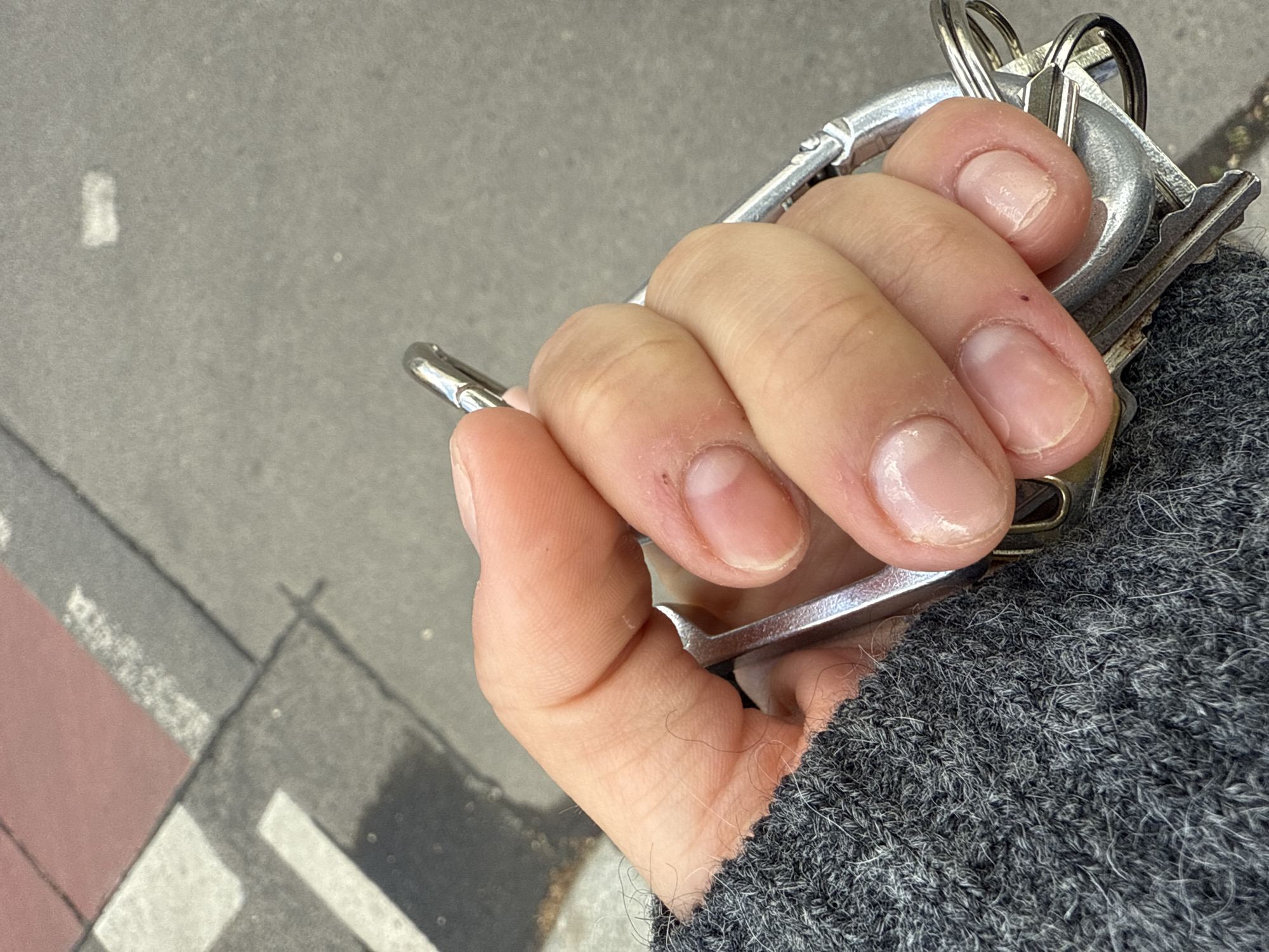 A hand holds a set of keys attached to a silver carabiner. The person is wearing a grey sweater. The background shows a street scene, identifiable as being in Berlin, Germany due to the presence of the red crosswalk lanes that are unique to Berlin.
