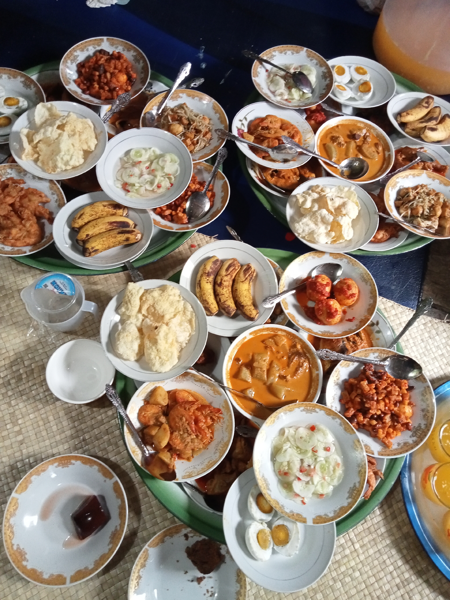 An overhead shot captures a spread of various Indonesian dishes arranged on a woven mat. The dishes are served in white bowls and plates with decorative gold rims. In the center, a bowl of orange curry with chunks of meat and vegetables is prominent, with a spoon resting inside. To the left, a bowl of white crackers sits next to a small pitcher of water. Other dishes include a portion of fried food, sliced bananas, a salad of cucumbers and red chilies, salted eggs, and a small cube of dark jelly or dessert. Several spoons are placed in or near the dishes, indicating they are ready for consumption. The background is dark, suggesting an indoor setting. The scene appears to be a meal preparation or a celebratory gathering.