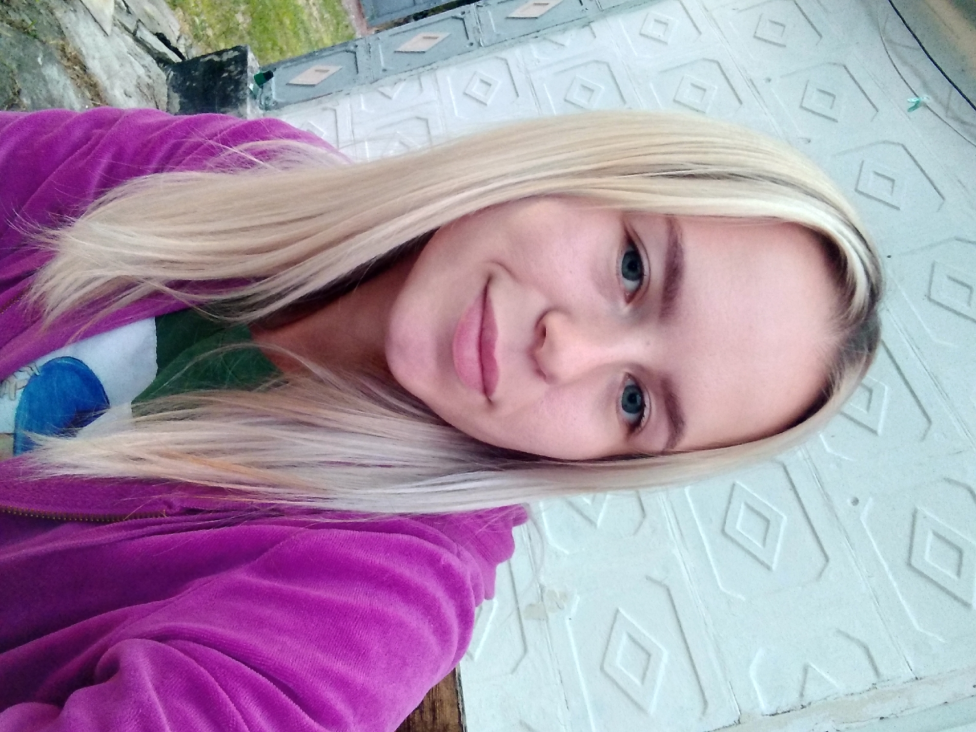 A young Caucasian woman with long blonde hair and blue eyes is pictured from the chest up, looking directly at the camera. She is wearing a bright purple velour hoodie over a green t-shirt. The background consists of a light-colored, textured wall with a diamond pattern. The setting appears to be outdoors, with a hint of greenery and rocks visible on the left side of the frame, suggesting a location in or near Radisne, Ukraine. The woman has a neutral expression with a slight smile.