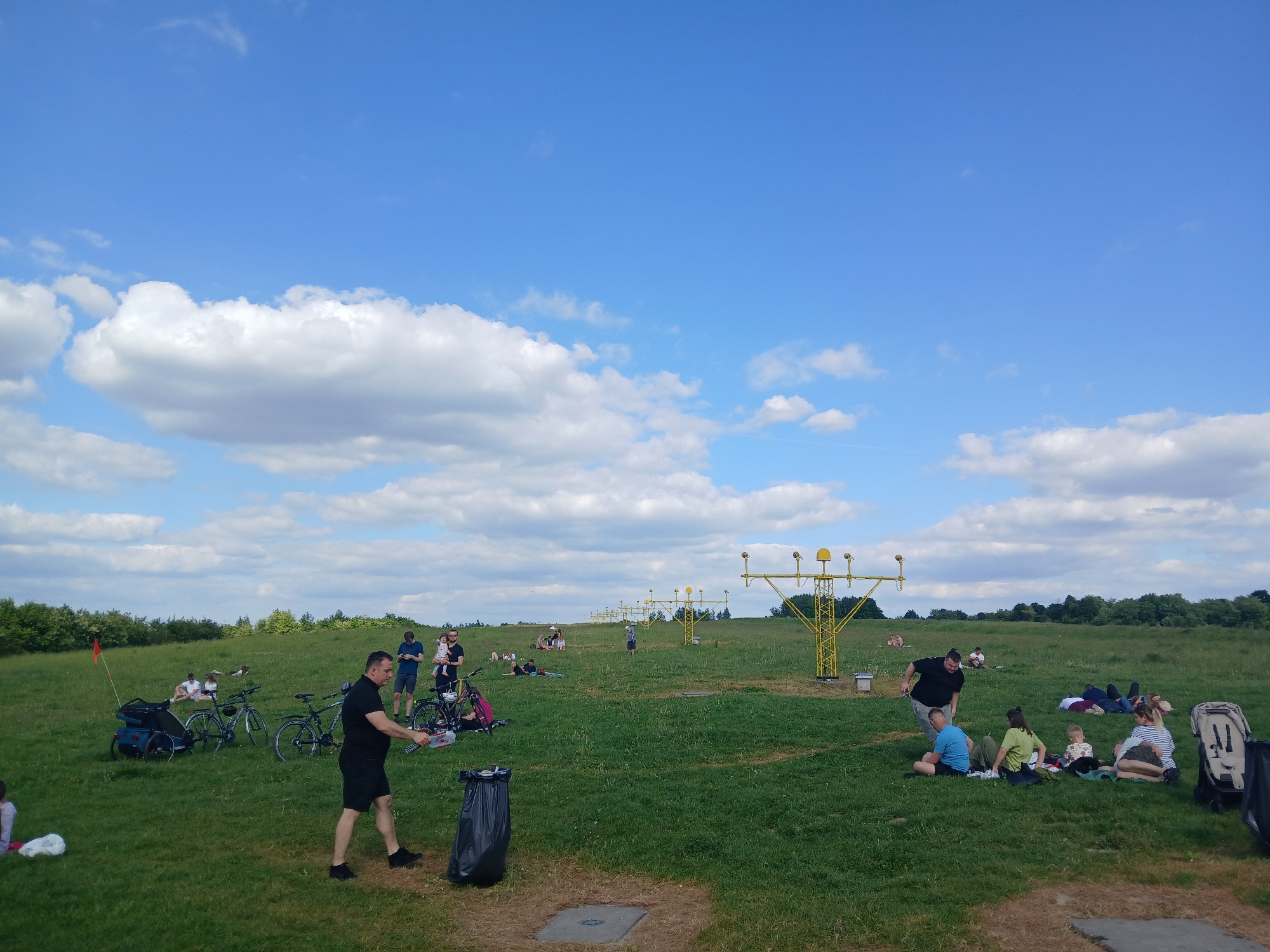 The image depicts a grassy field on a partly cloudy day in Unknown, Unknown. Numerous people are spread across the field, engaging in various activities. In the foreground, a man is seen throwing trash into a black bin. Several bicycles are parked nearby, one with a child's trailer attached and a red flag. In the midground, groups of people are picnicking and relaxing on the grass. A series of yellow metal structures with lights are visible in the middle of the field, spaced out at regular intervals. In the background, a line of trees borders the field. The sky is blue with puffy white clouds, taking up a significant portion of the image.
