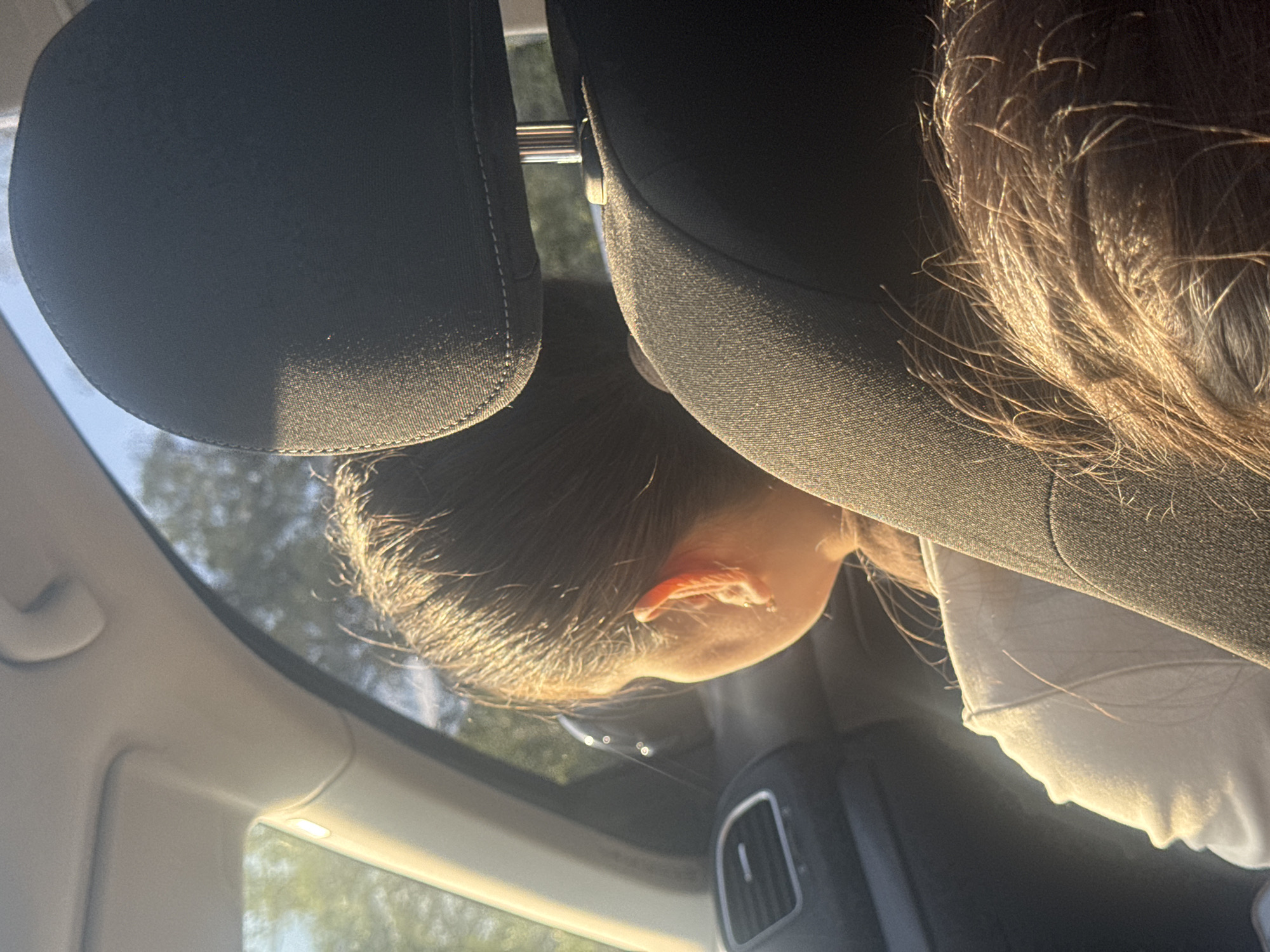This image captures the interior of an automobile in Suprunivka, Ukraine, viewed from a rear-passenger perspective. Two individuals are present within the vehicle. In the middle ground, a person with dark, pulled-back hair, wearing a light-colored long-sleeved top, is seated and looking out the right-side window. A dark fabric seatbelt diagonally crosses their chest and shoulder. A small, circular, metallic earring is visible in their right earlobe. In the lower right foreground, a mass of disheveled dark hair with prominent golden highlights from direct sunlight is visible, belonging to a second individual likely seated in front of the first person. Car interior components are visible, including a light-colored headliner, a dark dashboard with a silver-trimmed air vent, dark fabric seat upholstery, and a chrome headrest support post. Outside the vehicle, through the window, diffused green foliage and trees are visible against a bright sky, indicating an outdoor environment under direct sunlight. The scene is strongly illuminated by warm, golden natural light entering from the right, highlighting textures and surfaces within the car.
