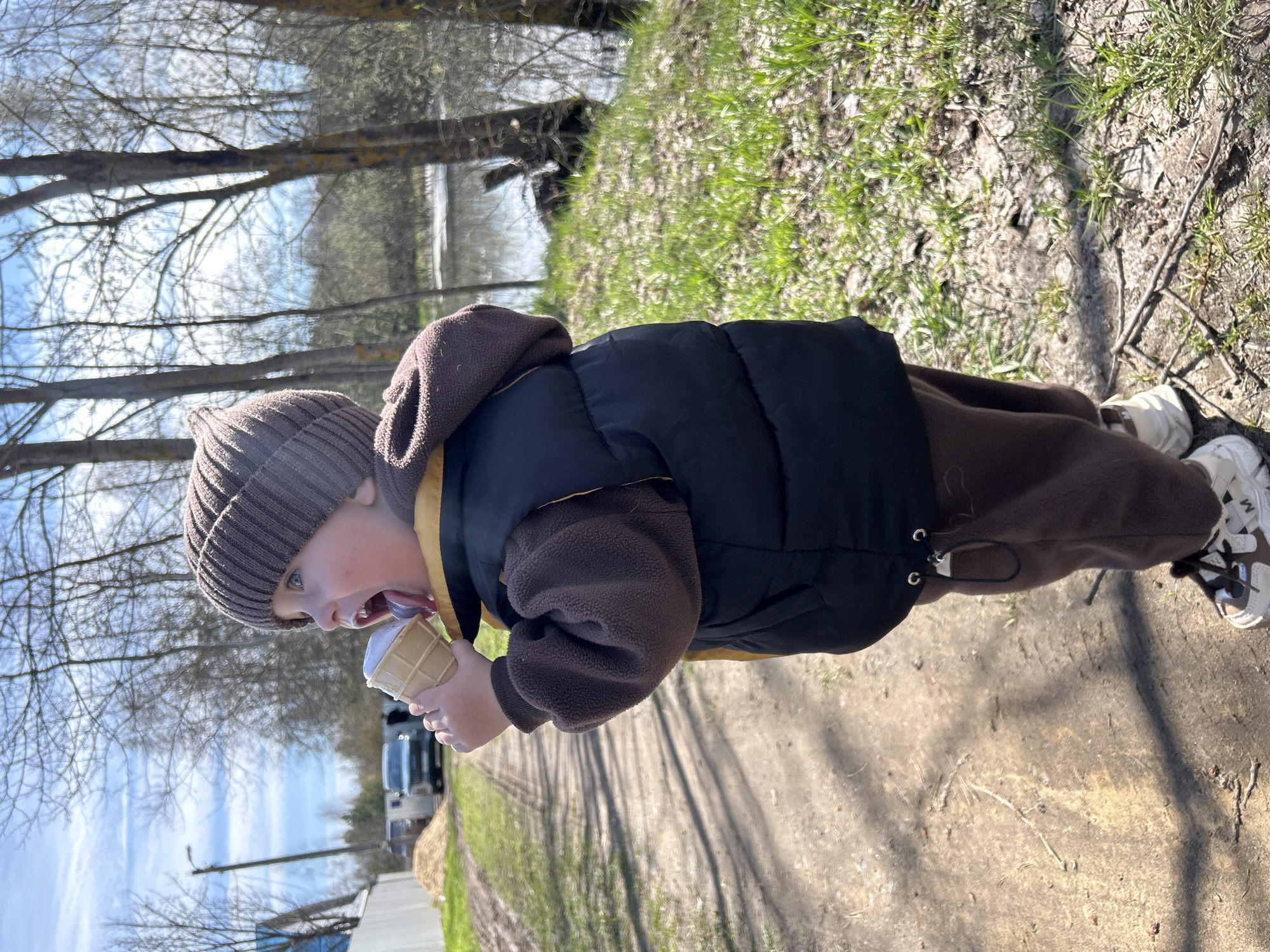 A young child, likely a toddler, is seen eating an ice cream cone while standing on a dirt path. The child is wearing a brown knit hat, a brown fleece jacket, and a black puffer vest. They also have on brown pants and white sneakers. The background shows trees with bare branches, suggesting early spring. To the left, there are some buildings and what appears to be parked vehicles. To the right, there is a grassy area. Based on the location, the scene is likely set in a rural or suburban area of Мещёры, Russia.