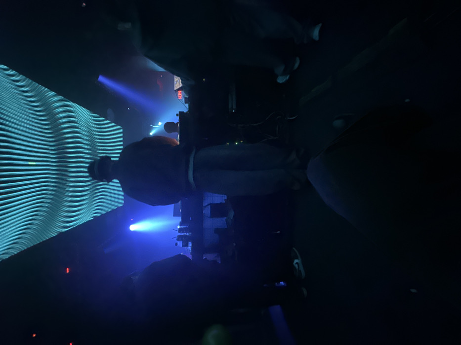 A nightclub scene, possibly a concert, features a person facing a large wavy LED screen bathed in blue light. Other patrons are indistinct in the dim environment. A stage is present, with equipment and lighting focused in the center. The atmosphere is dark and energetic, capturing a moment of performance or entertainment.