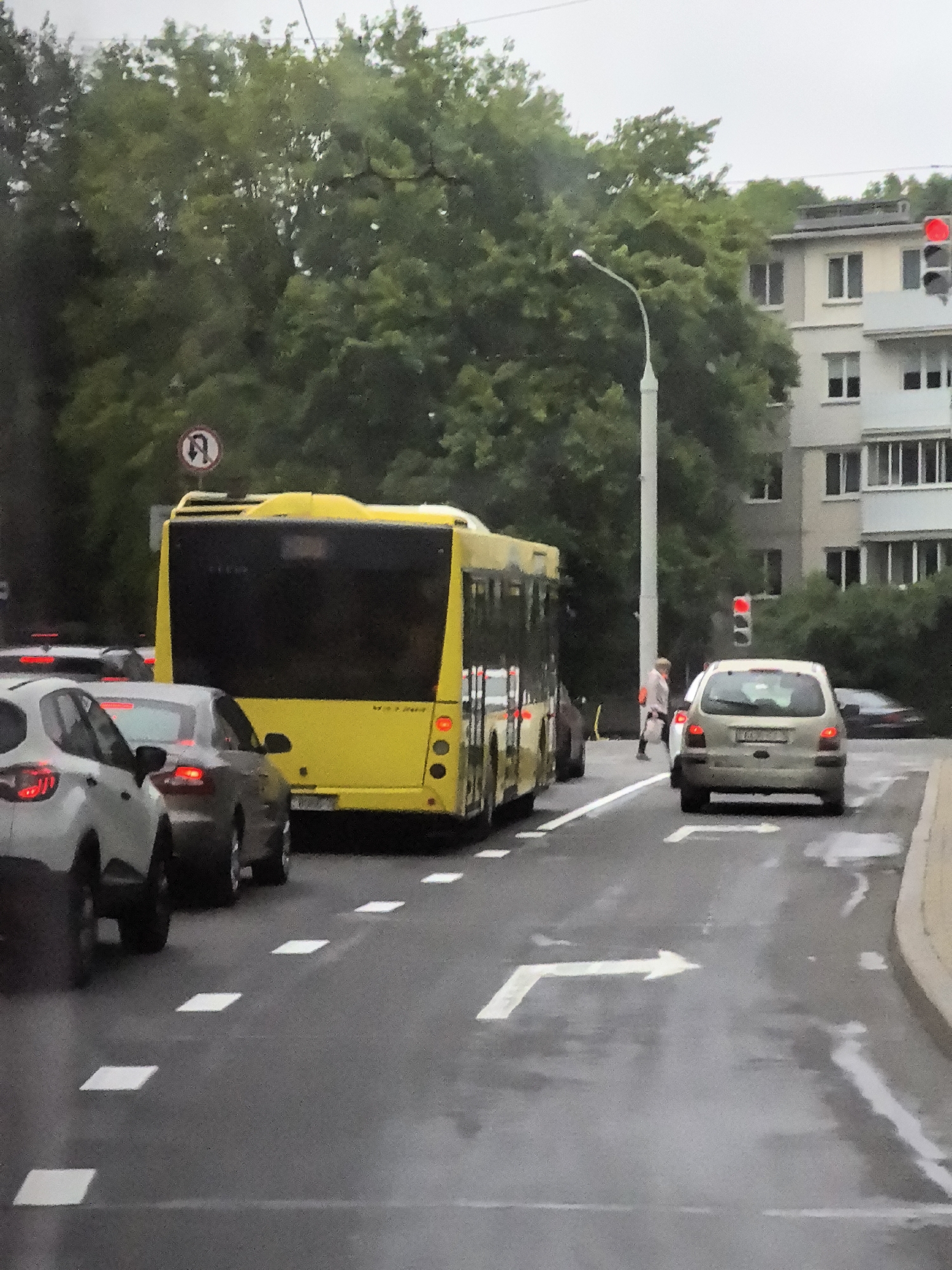 The image depicts a street scene in a city, likely on an overcast or rainy day, given the wet road surface and muted lighting. A bright yellow bus is the most prominent object, positioned in the center of the frame, facing away from the viewer. It has a large, dark rear window and several circular taillights. To the left of the bus, a silver car is partially visible, its taillights illuminated. Further left, another silver car is more fully in view, also with illuminated taillights. In the foreground, white lane markings, including arrows indicating turning directions, are visible on the asphalt. On the right side of the road, a light-colored compact car is stopped, its brake lights on. Behind this car, a building with multiple windows and balconies rises. A pedestrian, a person with blonde hair wearing a light-colored jacket and dark pants, is walking on the sidewalk or road near the pedestrian crossing, partially obscured by a traffic light pole. A street lamp is also visible. In the background, lush green trees line the street. A traffic sign with a red circle and a diagonal line across a curved arrow is visible on the left side of the road, indicating a prohibition against a U-turn. A traffic light with a red signal is visible on the right side of the road.