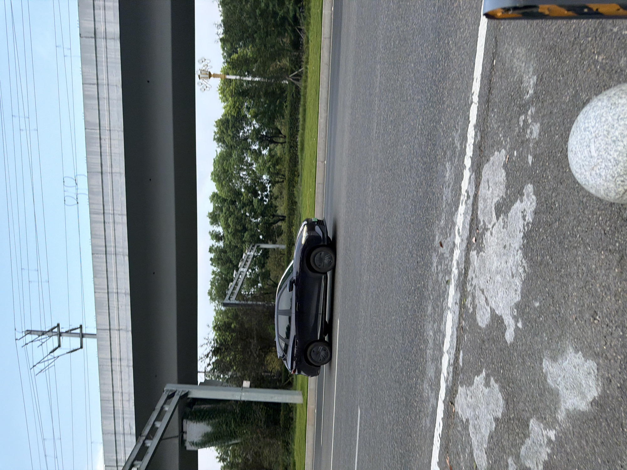 A dark-colored, modern SUV is driving on a paved road in Jiangjin, China. The vehicle, appearing to be in motion from left to right, is positioned in the right lane of the road, which has white lane markings. In the background, a multi-tiered elevated structure, likely part of a bridge or overpass, looms above. Power lines and transmission towers are visible against a light blue sky in the upper portion of the image. Further in the background, a line of lush green trees and vegetation runs along the side of the road. To the right of the road, a decorative lamppost with multiple lights stands. The foreground shows the textured asphalt of the road surface with some lighter patches, possibly from wear or repairs, and a white line marking the edge of a lane. A round, grey, stone bollard is visible in the lower right corner of the frame.