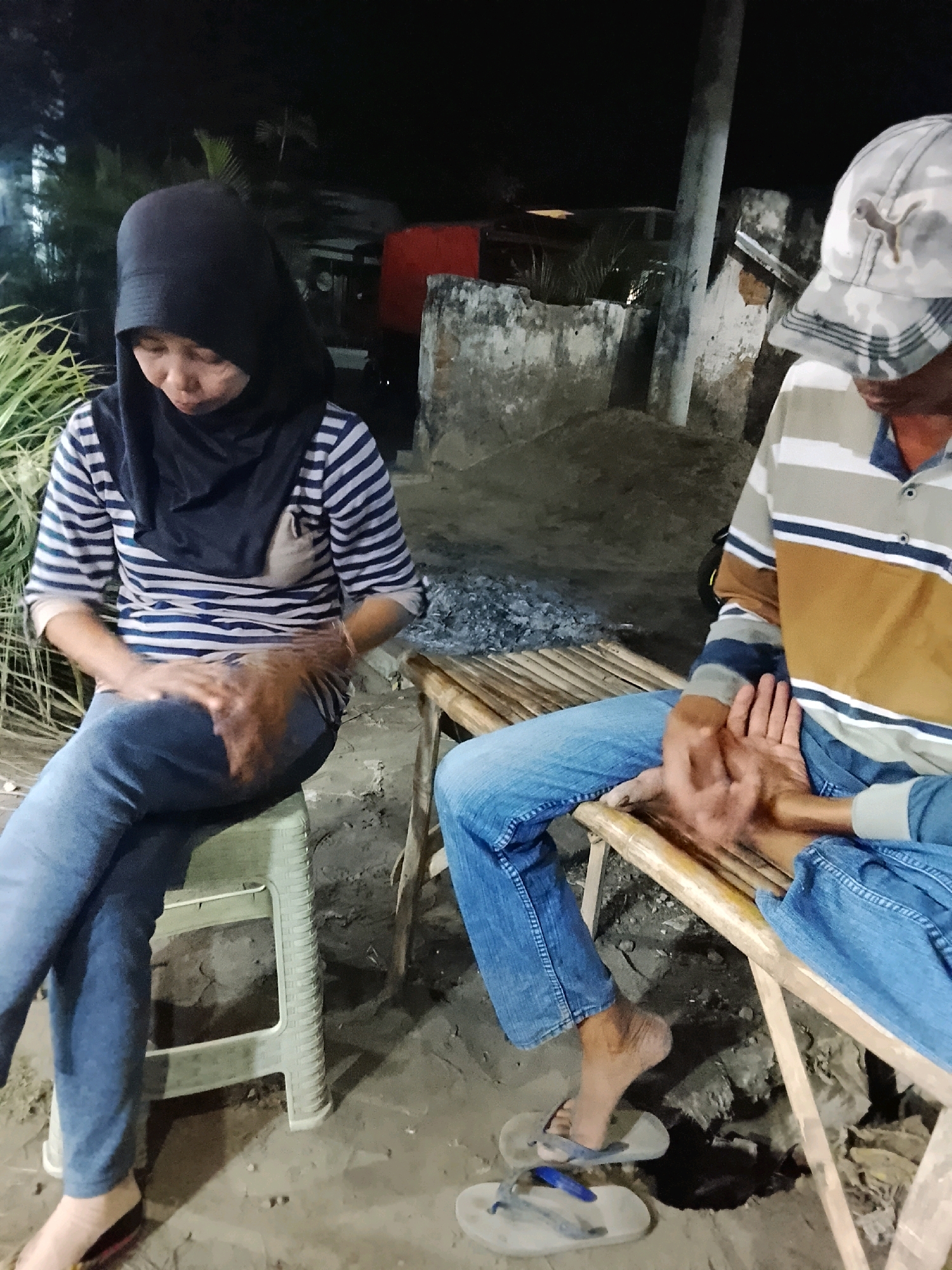 In Sukorejo, Indonesia, at night, two individuals are seated outdoors on a sandy ground. A woman wearing a black hijab and a horizontally striped long-sleeved shirt sits on a green plastic stool. Her hands are clasped in her lap. To her right, a man wearing a camouflage baseball cap and a horizontally striped polo shirt is seated on a bamboo bench. His hands are clasped together. The environment is dimly lit, with some foliage visible in the background, along with a concrete wall and a pole. The ground appears to be dirt and sand, with some scattered debris.