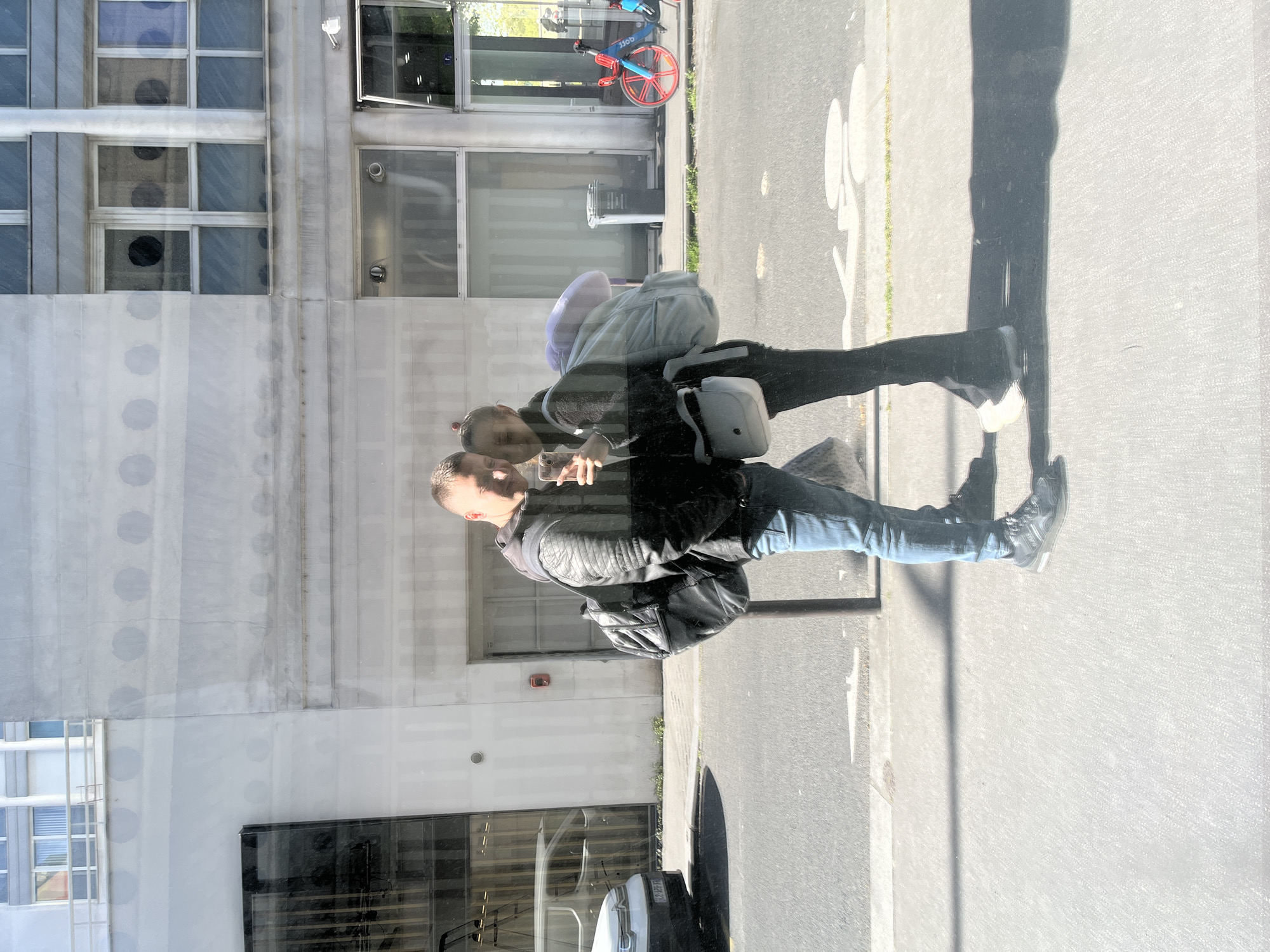 The image presents a complex urban scene in Paris, captured through a highly reflective glass surface, likely a building's window or facade. The primary subjects are the reflections of two individuals. On the left, a man with short hair, dressed in a dark leather jacket over a hooded sweatshirt and blue jeans, holds a smartphone to his face, appearing to take a selfie or video. He wears a dark backpack. Beside him and slightly behind, a woman with long hair tied back, wearing a light-colored top and dark trousers, has her arm around the man. She carries a light shoulder bag. Their reflections indicate a close, possibly affectionate, interaction.
Beyond the reflections, elements of a bustling street and a modern building are visible. The pavement consists of asphalt and a concrete sidewalk, marked with white painted road symbols, including a bicycle icon and an arrow. A street light pole is partially obscured by the reflected figures. In the background, a blue and red shared bicycle, bearing the text "jCD" (identifying it as a JCDecaux bike, common in Paris), is parked near a building entrance. The building itself is a multi-story structure with numerous large windows, some revealing interior details like circular lights and open windows.
The scene is illuminated by bright natural sunlight, creating distinct, long shadows on the pavement, suggesting a sunny late morning or early afternoon. The interplay of reflections and transparency adds depth to the urban landscape. The specific "jCD" bicycle text and the overall urban architecture are strong indicators of the Paris setting. A white car's front bumper and a partially visible license plate ("AZ-129-YA") are seen at the bottom right.
