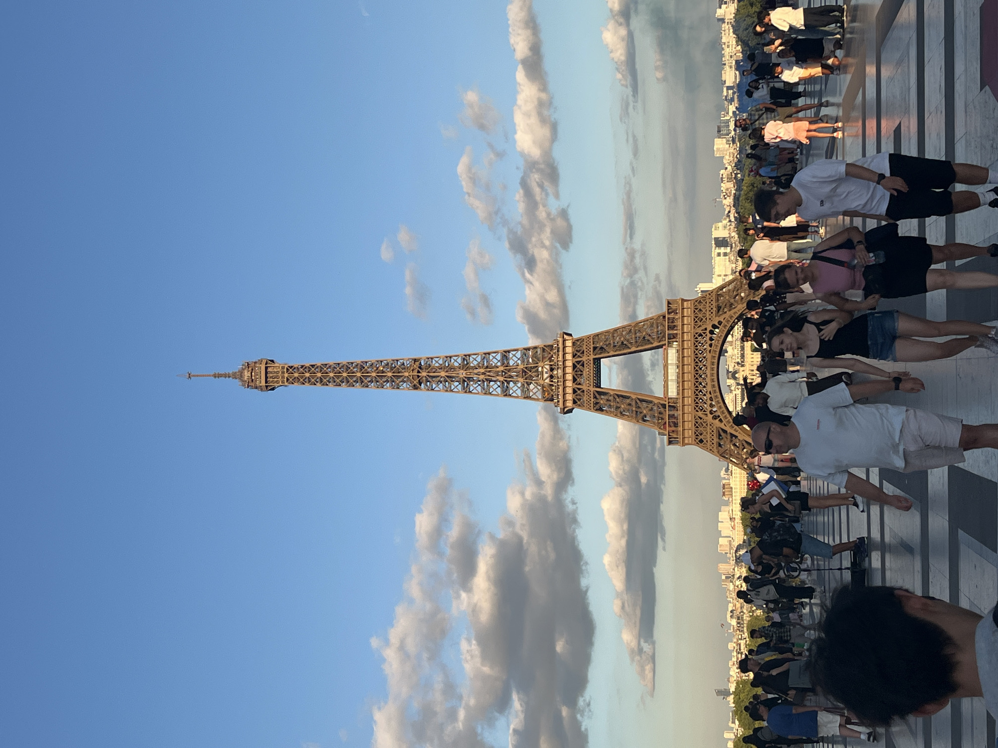 The image shows the Eiffel Tower in Paris, France, under a clear blue sky with scattered clouds. In the foreground, a crowd of tourists is visible, some walking and others standing. The Eiffel Tower is the central and dominant subject, its intricate iron structure clearly defined against the sky. The area around the tower appears to be a public plaza with a patterned pavement. In the background, the Parisian cityscape is visible with buildings stretching towards the horizon. The lighting suggests it is late afternoon or early evening.