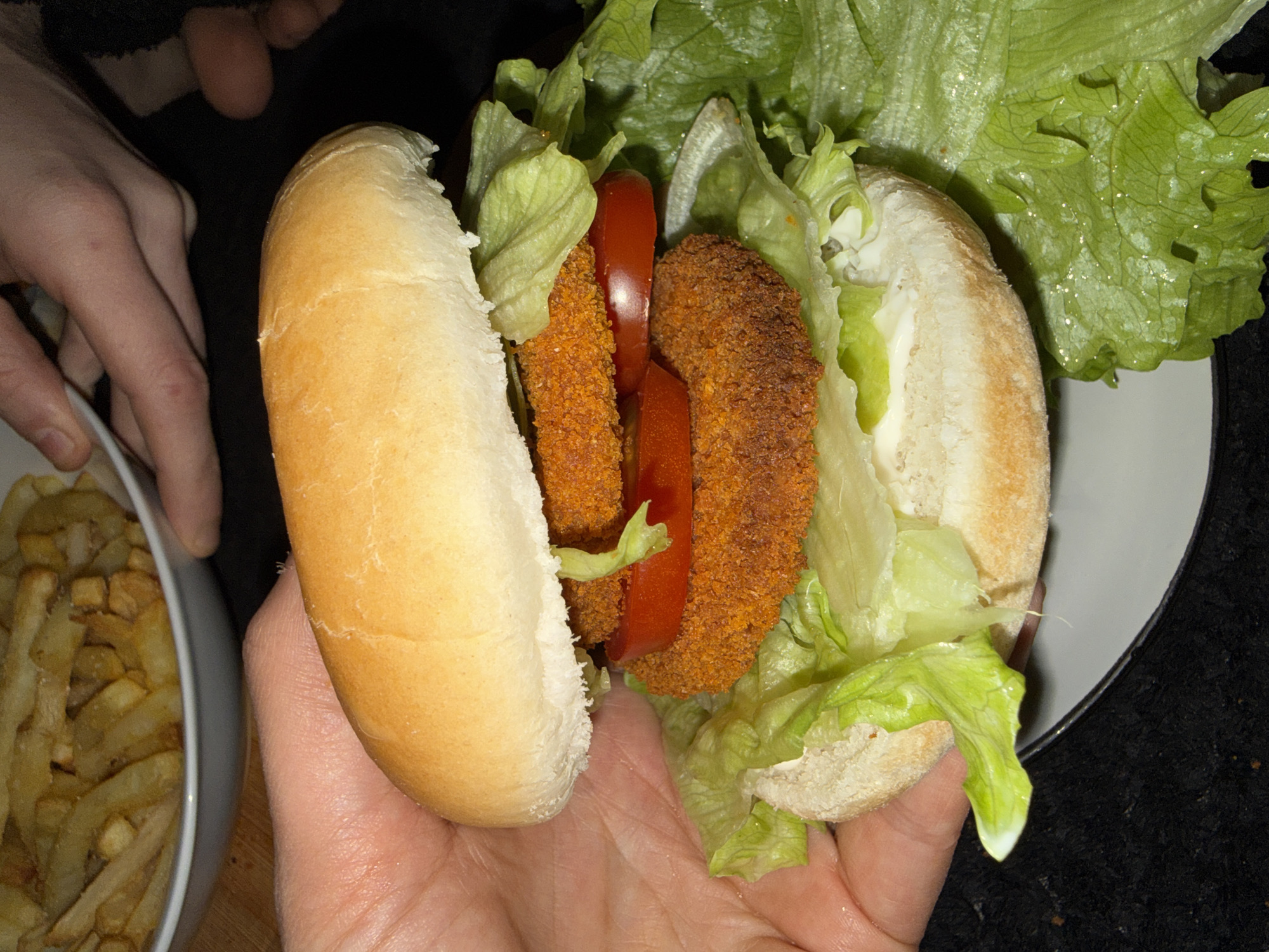A person holds a chicken fillet sandwich in their hand. The sandwich consists of a bun, two breaded chicken fillets, lettuce, and sliced tomatoes. A bowl of French fries is visible in the background to the left. The location is not discernible from the image. The city of Great Yarmouth, United Kingdom, is also not discernible.