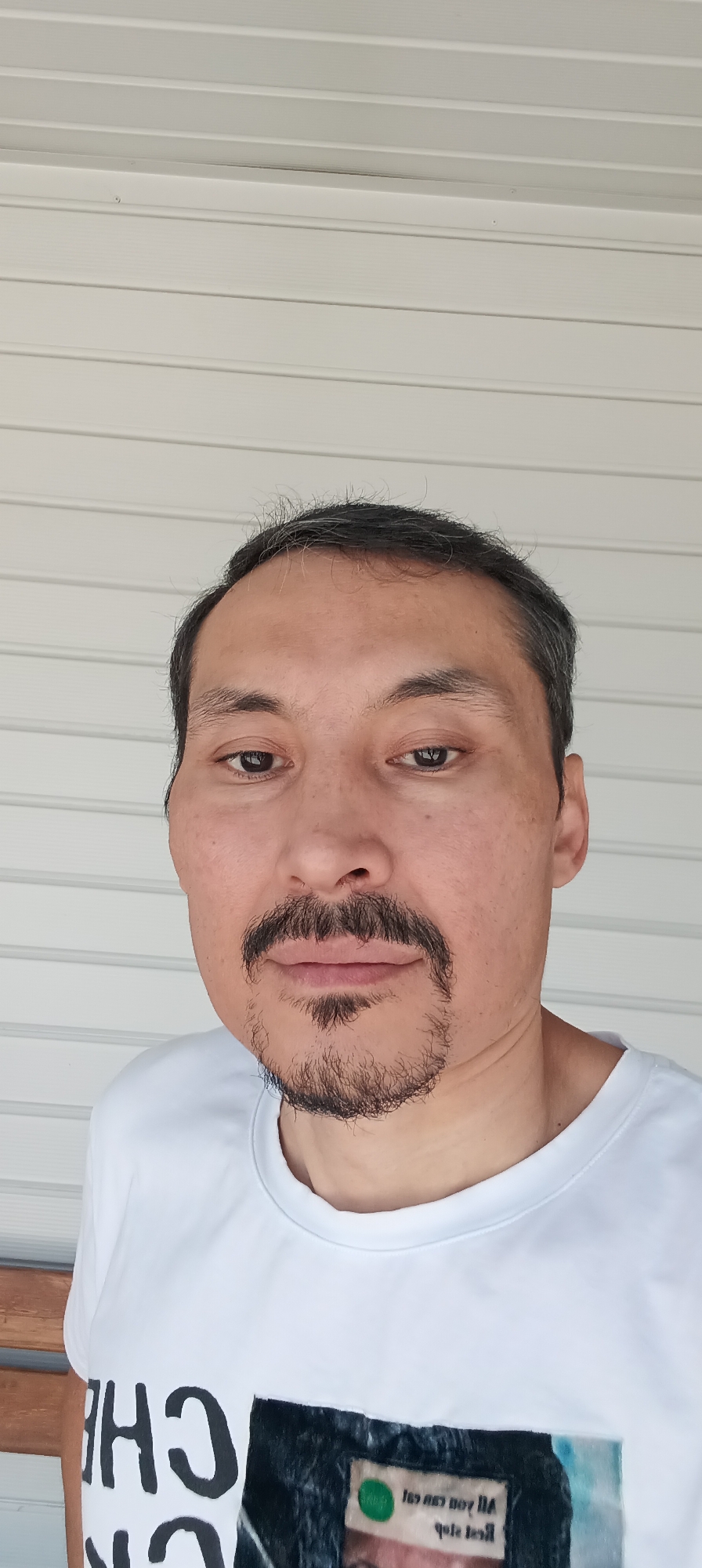 A man with dark hair and a mustache and goatee is shown from the chest up, looking at the camera. He is wearing a white t-shirt that has text and an image on the front. The background is a white, horizontal slatted surface, possibly siding on a building. The image appears to be taken outdoors during the day. There is no discernible indication of the location in the city of Qarağandı city administration, Kazakhstan.