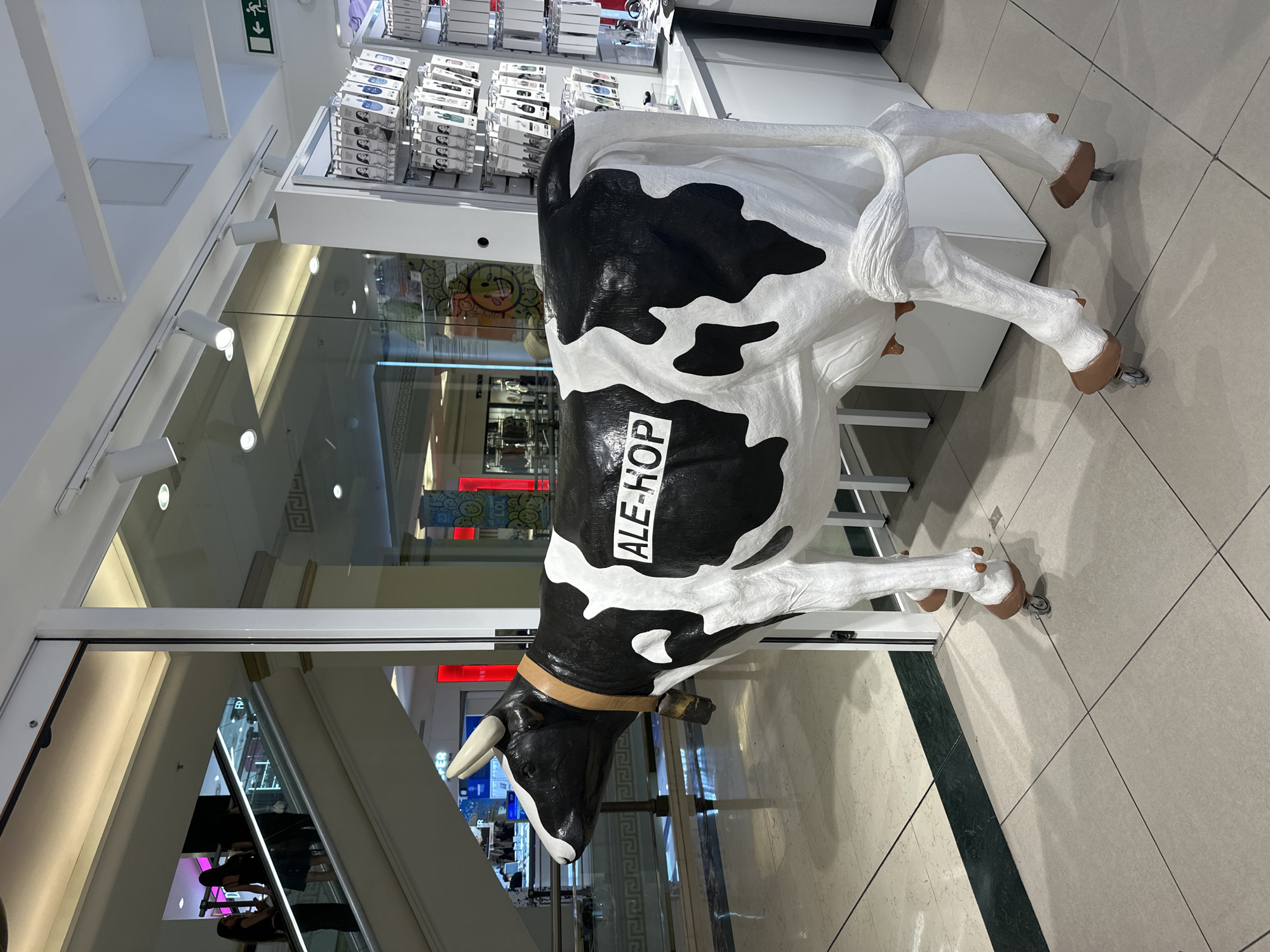 A life-size black and white cow statue with wheels on its hooves stands on a tiled floor inside a brightly lit store. The cow has a tan collar with a bell and a white rectangular sign on its flank that reads "ALE-HOP". Behind the cow, a glass-paneled store front shows escalators and shoppers in the background, suggesting an indoor shopping mall environment. To the right of the cow, shelves stocked with small electronic accessories are visible.