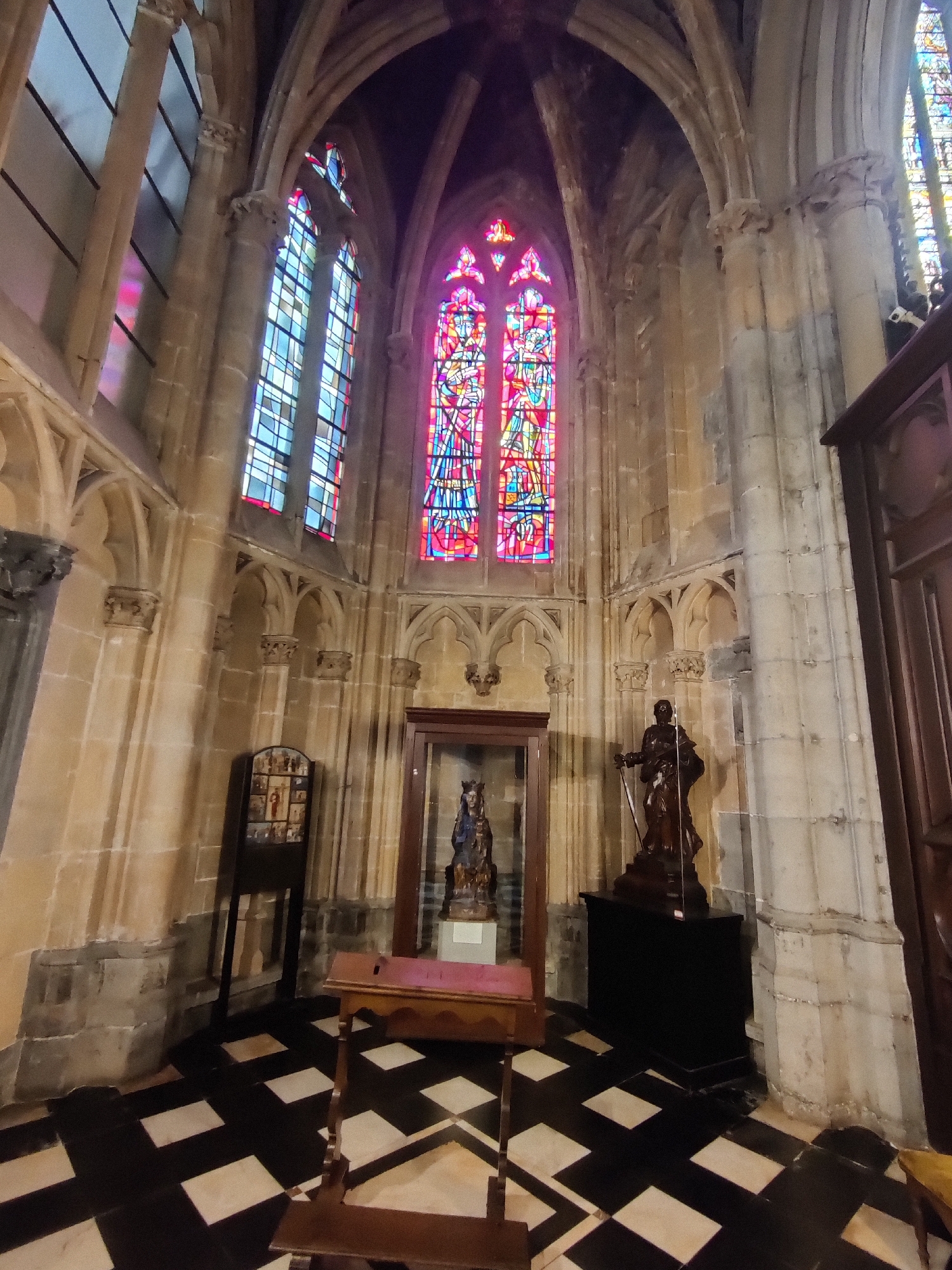 The image shows the interior of Saint Paul's Cathedral in Liège, Belgium, specifically the choir area, featuring a large stained-glass window with red and blue patterns. In the foreground, a black and white checkered tile floor is visible. To the left, a wooden stand displays a series of small religious icons. In the center, a glass display case contains a statue of the Virgin Mary. To the right, a dark statue of a robed figure holding a sword stands on a dark pedestal. The architectural style suggests Gothic influence, with stone columns and arched ceilings. The lighting is dim, with the stained glass being the primary source of colorful illumination.