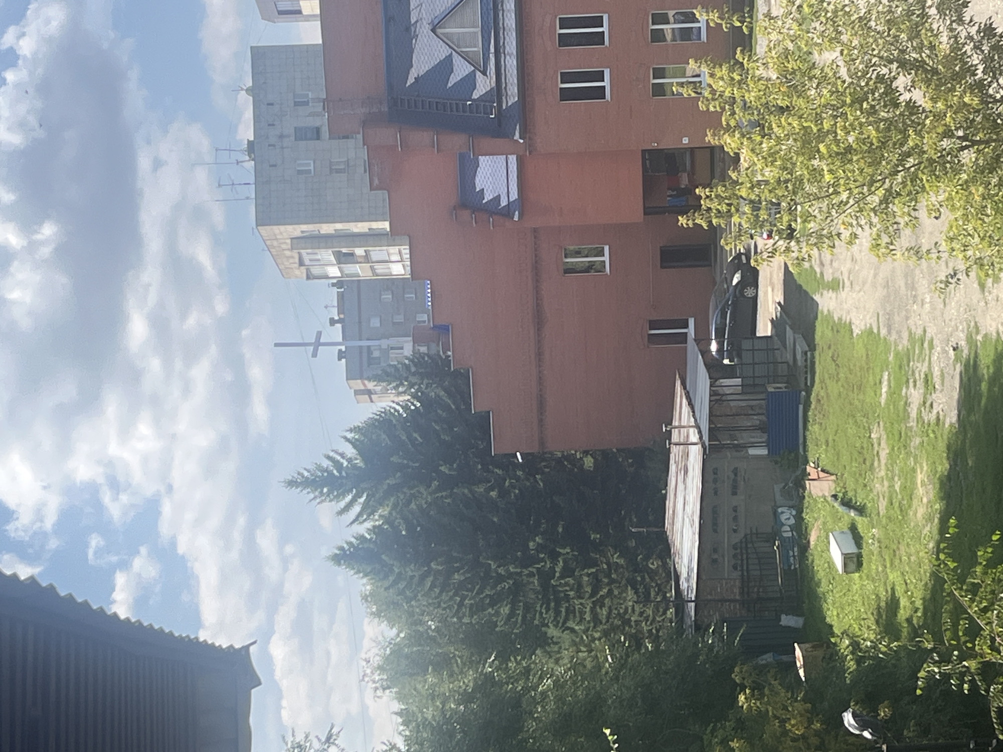 The image captures a view of a multi-story brick building with a dark blue roof against a partly cloudy sky. The building features numerous windows and a prominent staircase on its exterior. In front of this building, there is a smaller, single-story structure with a corrugated metal roof, surrounded by green grass and some scattered objects including a white rectangular container. Several trees, some appearing to be coniferous, are visible in the middle ground and to the left of the frame. In the background, several taller apartment buildings can be seen, some with what appear to be antennas on their roofs, and a white cross structure. A black car is parked on a paved area in front of the brick building, next to the smaller structure. The scene appears to be an outdoor area in a residential or mixed-use urban environment, possibly in the city of Oskemen, Kazakhstan, given the architectural styles and vegetation.