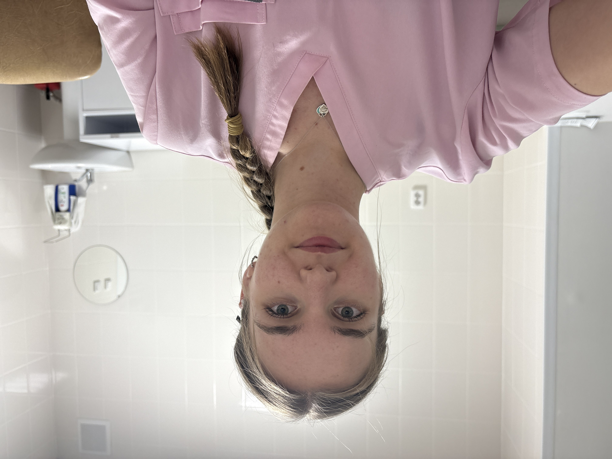 A young woman with blonde hair braided and pulled to one side is posing for a selfie. She wears a pink medical scrub top and a silver necklace. She is standing in what appears to be a tiled medical or dental examination room with a dental chair and equipment in the background. The location is possibly in Perm, Russia.