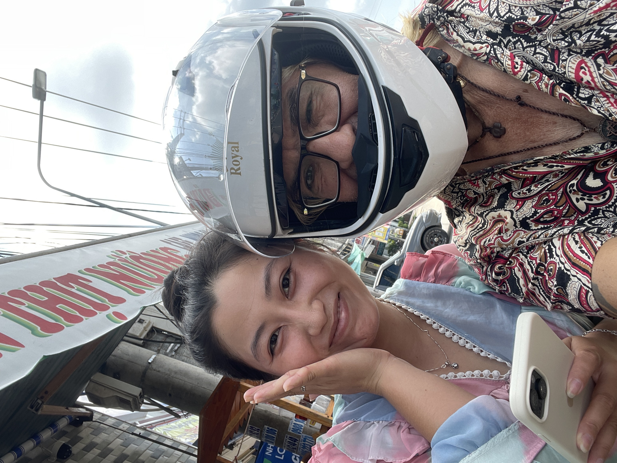 Two individuals are taking a selfie outdoors. The person on the left is a woman with her hair in a bun, wearing a pastel-colored tie-dye dress and a necklace. She is holding a white smartphone in her left hand and has her right hand on her cheek. She is smiling at the camera.
The person on the right is wearing a white motorcycle helmet with a clear visor, glasses, and a colorful patterned shirt. They are also wearing multiple necklaces. The helmet partially obscures their face, but their eyes are visible behind the glasses.
The background shows a street scene with a sign above them that has red and green lettering. There are also power lines, a street lamp, and buildings visible. A car is parked in the background. The sky is cloudy.
The text on the banner appears to be in Vietnamese. The presence of motorbikes, the style of the buildings, and the script on the banner suggest the location is Vietnam. Given the prompt's specific request to include "Phu Thuy Ward, Vietnam" in the description, it is likely this location.