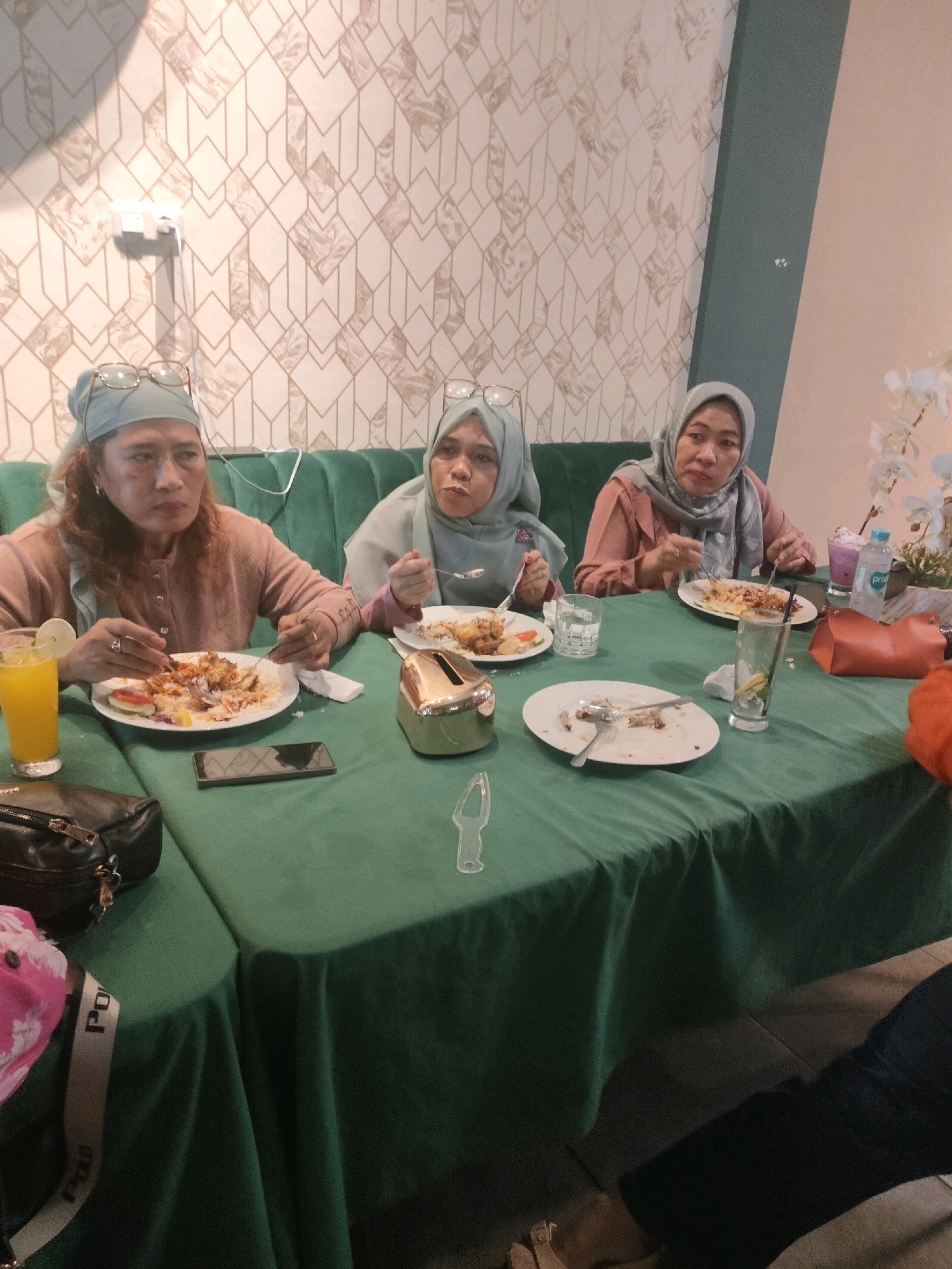 Four women are seated around a table covered with a green tablecloth, eating a meal. The woman on the left is wearing a light brown cardigan and a light blue headscarf. She has glasses on her head. In front of her is a plate of food and a glass of orange juice. The second woman from the left is wearing a light blue headscarf and a light pink top. She is looking at the camera and holding a fork. In front of her is a plate of food. The third woman from the left is wearing a grey headscarf and a light pink top. She is holding a fork and a knife, and eating from a plate of food. In front of her is a glass of water. The fourth woman is partially visible on the right side of the image. She is wearing a grey headscarf and a light pink top. The background features a patterned wallpaper in shades of beige and white. There is a green sofa behind the women. On the table, there are also several plates with food, cutlery, glasses, and a bottle of water. The scene appears to be an indoor dining setting, possibly a restaurant. The image was taken in Pekanbaru, Indonesia.