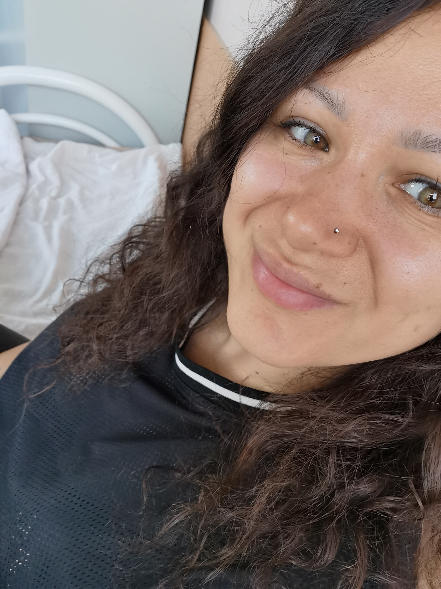 A woman with brown curly hair and a nose piercing is looking towards the upper right corner of the frame while smiling. She is wearing a black athletic shirt with a white stripe at the neckline. In the background, there is a white headboard and a white sheet. The image does not provide sufficient information to determine the location in Perm, Russia or any actions, interactions or further environmental details.