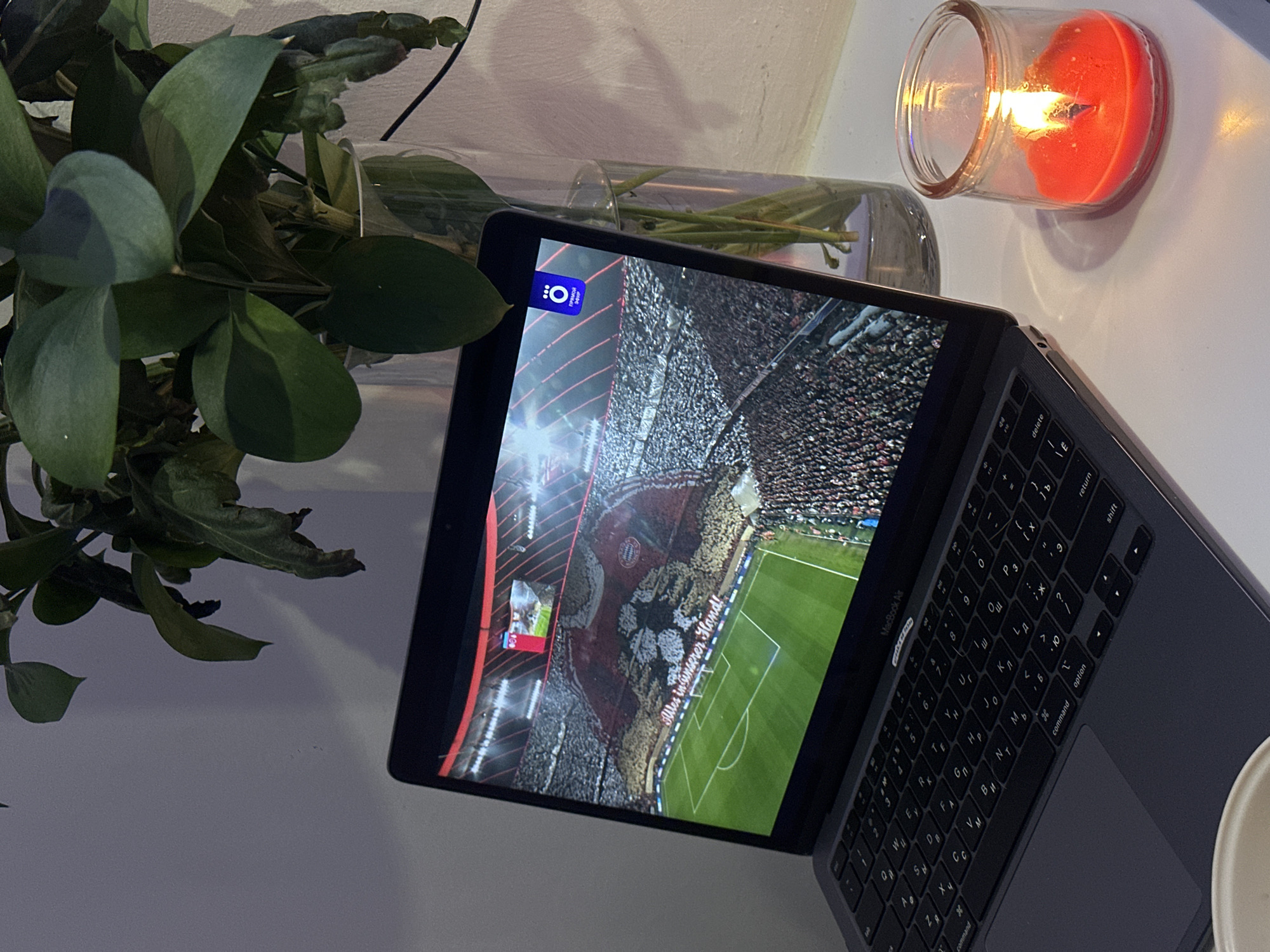 Here's a detailed description of the image:
**Scene Overview:** The image captures a still life arrangement on a table or desk. Key elements include a laptop displaying a soccer game, a plant in a vase, and a lit candle.
**Objects:**
* **Laptop:** A "MacBook Air" laptop occupies the center of the frame. The screen displays a soccer game, showing a view of the field, players, and spectators in the stands. The image includes the visible portion of the keyboard.
* **Plant:** A leafy green plant is placed to the left of the laptop, with some leaves overlapping the edge of the screen. The plant is in a clear glass vase filled with water.
* **Candle:** A small, lit candle in a glass container is located to the right of the laptop, near the edge of the table. The wax appears red/orange.
**Environment:**
* The setting appears to be indoors, possibly in a home or office space.
* The surface the objects rest on appears to be white.
**Implied Context:** Based on the image content, it's plausible the person in the setting is watching a soccer game on their laptop, possibly in a relaxed or cozy environment (indicated by the candle).
**Location:** The keyboard appears to have both Latin and Cyrillic lettering, which suggest that the location is in Russia. Given the language of "Mes in unserer Hand!" displayed on the laptop screen, and the depiction of the Allianz Arena, it's very likely a match of Bayern Munich. Therefore, the soccer game does not take place in Moscow.
