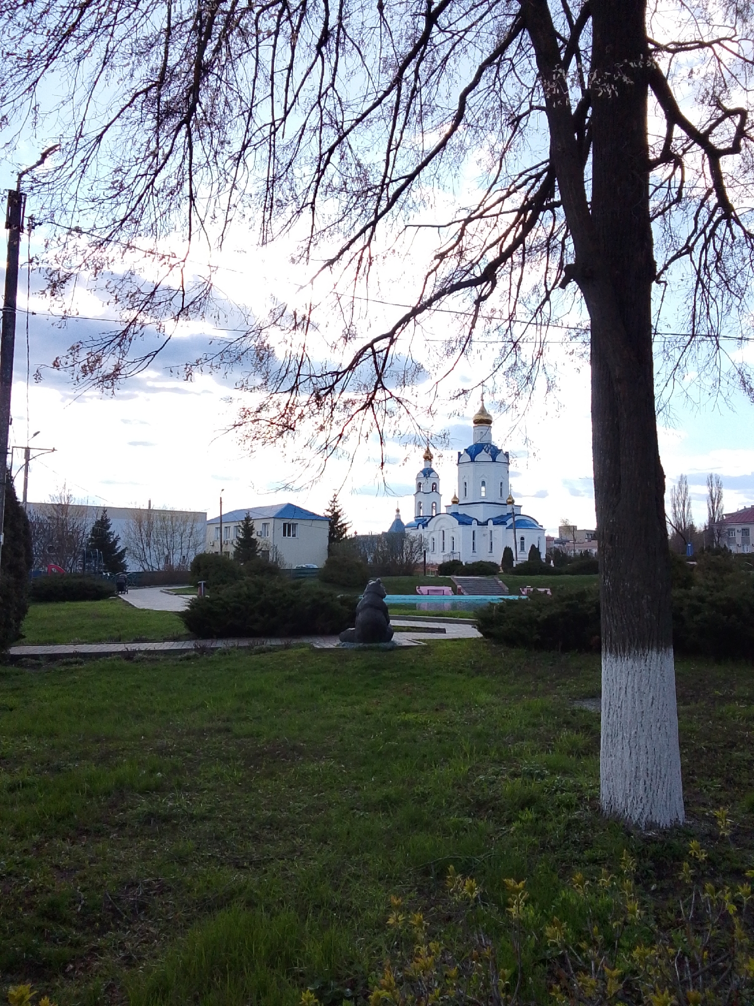 The image depicts an outdoor scene in Khlevnoye, Russia, featuring an Orthodox church in the background with white walls and blue roofs. The church has golden domes. In the foreground, there's a grassy area with bushes and trees. A bare tree trunk painted white at the base is prominently positioned on the right side of the frame. In the background left there is a flat roof building with a blue roof. A statue of a bear is also visible in the park. The sky is partly cloudy. The photograph is taken during the daytime.