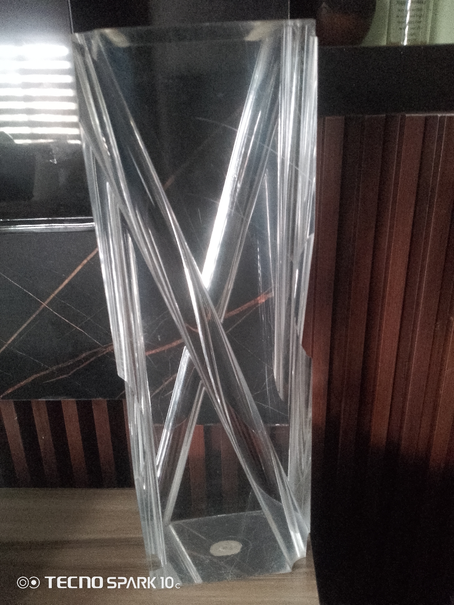 The image shows a clear, cylindrical glass vase with a twisted design, standing on a wooden surface. Behind the vase, there is a dark, marbled surface with thin, golden lines. To the right of the vase, a dark brown, ribbed structure is visible, possibly a piece of furniture or a wall. The background is dark and blurred, with some bright horizontal light patterns on the left, suggesting light coming through blinds or a similar aperture. The bottom left corner of the image contains the text "TECNO SPARK 10c" along with a logo, indicating the device used to capture the photograph. The scene appears to be indoors.