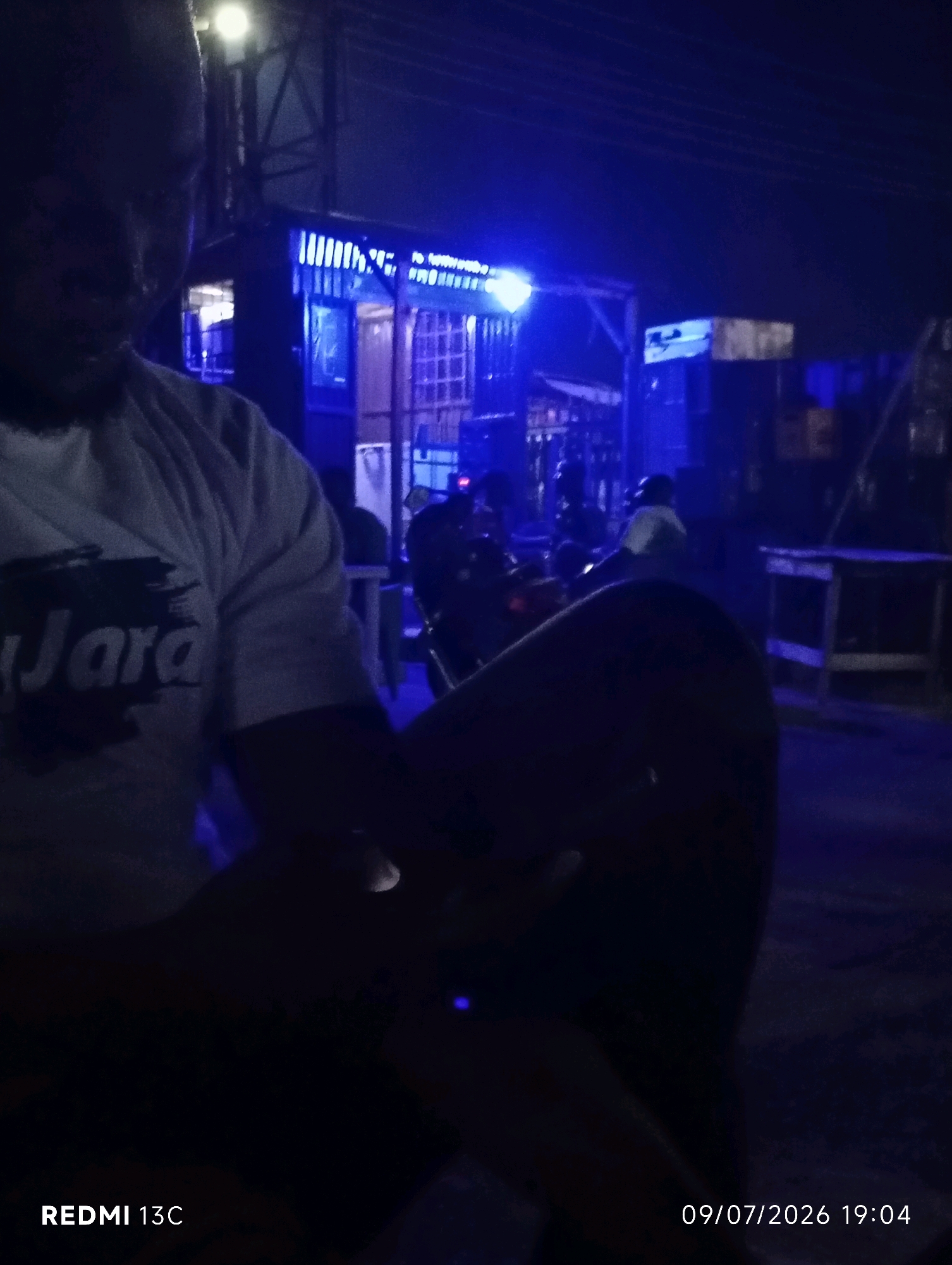 The image shows a close-up of a person in the foreground, appearing to be seated, with a dark blue and purple glow emanating from the background. The person is wearing a white t-shirt with a dark graphic that appears to say "Jara" in stylized lettering. The background depicts an outdoor scene at night, possibly a street or market area, with structures illuminated by blue lights. There are indications of other people and possibly motorcycles or vehicles in the distance. The overall atmosphere is dark and dimly lit, with the blue illumination creating a distinct nocturnal ambiance. The bottom right corner of the image displays a timestamp: "09/07/2026 19:04" and "REDMI 13C". This suggests the photo was taken in Shendam, Nigeria, on July 9, 2026, at 7:04 PM.