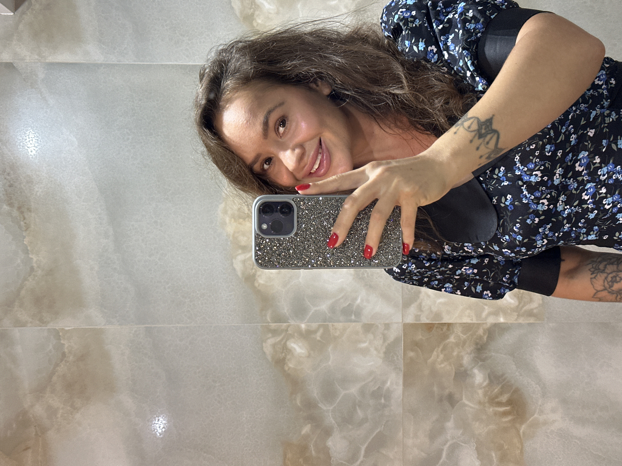 A woman is taking a selfie in a bathroom with marble-patterned walls. She is smiling and holding a phone with a sparkly case. She is wearing a black dress with a floral pattern. There is a tattoo on her arm.