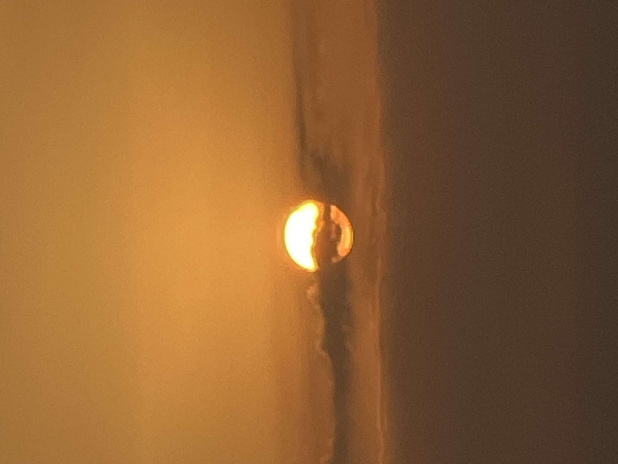 A full shot captures a sunset over Kuta, Indonesia. The sun, a glowing orb of orange and yellow, is partially obscured by a horizontal band of clouds. The sky above is a gradient of warm orange hues, while the sky below the clouds is a muted, hazy brown, possibly indicating the ocean or land shrouded in mist. The image is devoid of any people, man-made structures, or specific landmarks that would pinpoint a precise location within Kuta.