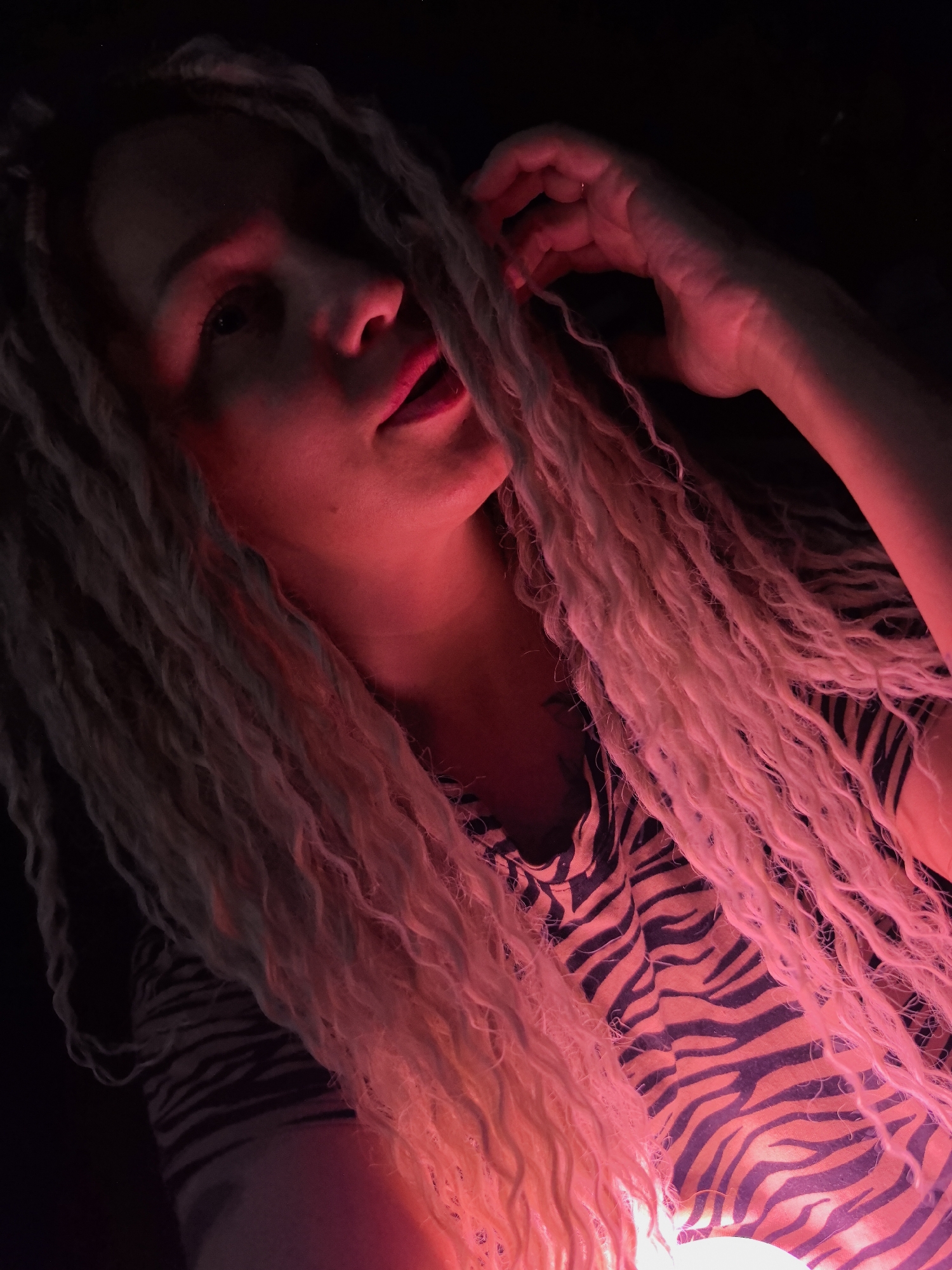 A person with light-colored dreadlocks is illuminated by a red light source in a dimly lit environment. The person is wearing a zebra-striped shirt and has their hand near their head, possibly touching the dreadlocks. The background is dark and undefined. There is no evidence of the location being in the city of Noviy, Russia.