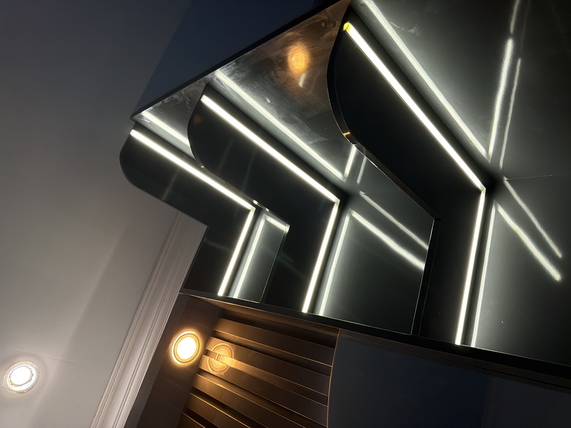 The image shows a section of a modern architectural design, likely an interior staircase or hallway, with a focus on sleek, dark surfaces and integrated LED lighting. The structure features sharp angles and curved elements, with multiple levels illuminated by long, thin white LED strips that create a futuristic and sophisticated ambiance. One section of the structure, to the left, has a darker, ribbed texture with a warm, spotlight-like illumination. The ceiling above is white with decorative molding and two recessed circular lights. The overall impression is one of contemporary luxury. There are no people visible in the image. The scene does not contain any specific information that would allow for its localization within Wonoayu, Indonesia, or any other specific city.